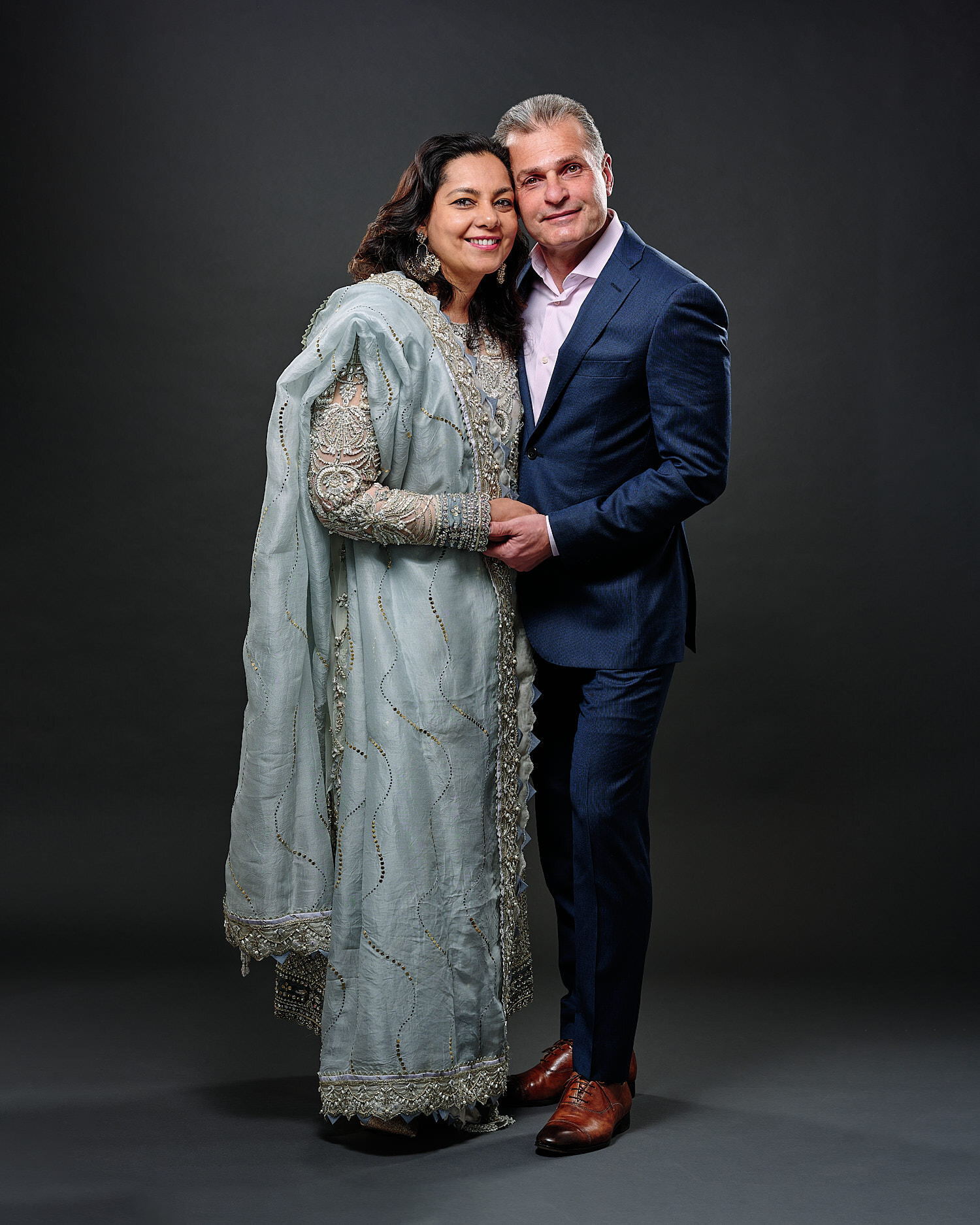  Bushra and Saad are posing for their wedding portraits in a professional photography studio. Bushra is dressed in a stunning pale dress ornamented with pearls, floral motives. Saad wears a blue suit. 