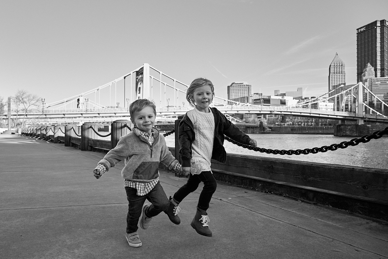  Crela Fry is posing with her husband and children by Allegheny River on the North Shore in Pittsburgh, Pennsylvania. Images are monochrome. The cityscape with yellow bridges is lit by the setting sun. 