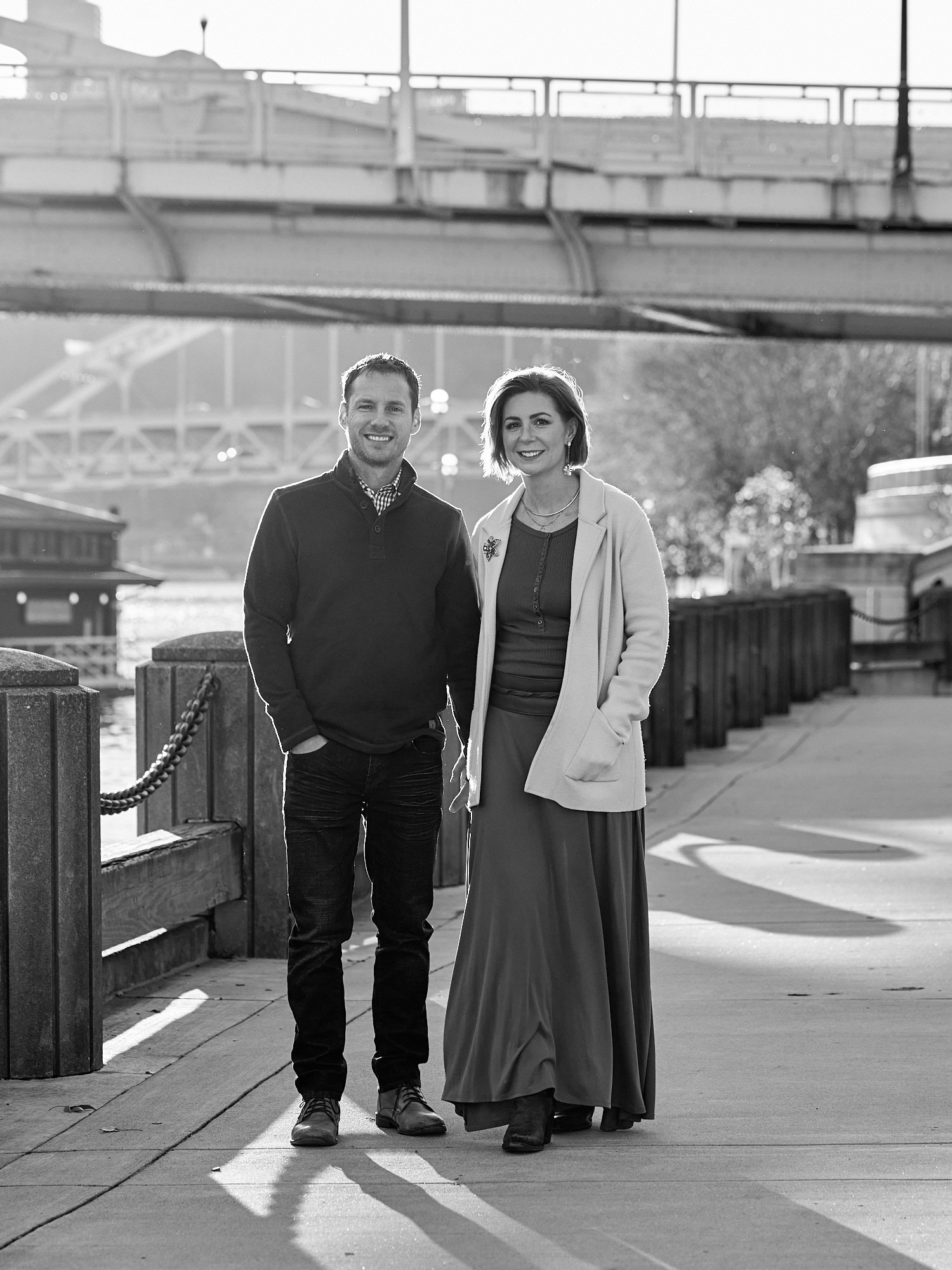  Crela Fry is posing with her husband and children by Allegheny River on the North Shore in Pittsburgh, Pennsylvania. Images are monochrome. The cityscape with yellow bridges is lit by the setting sun. 