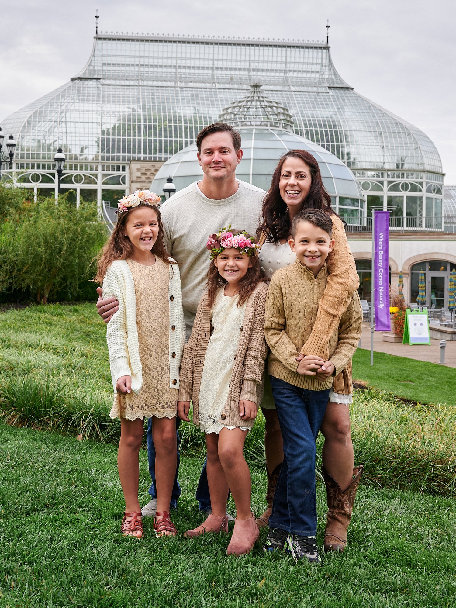  Zack Reed is posing with his wife, son and two daughters in front of Phipps Conservatory and Botanical Gardens of Pittsburgh, Pennsylvania. The ladies are wearing embroidered beige dresses and flowers 