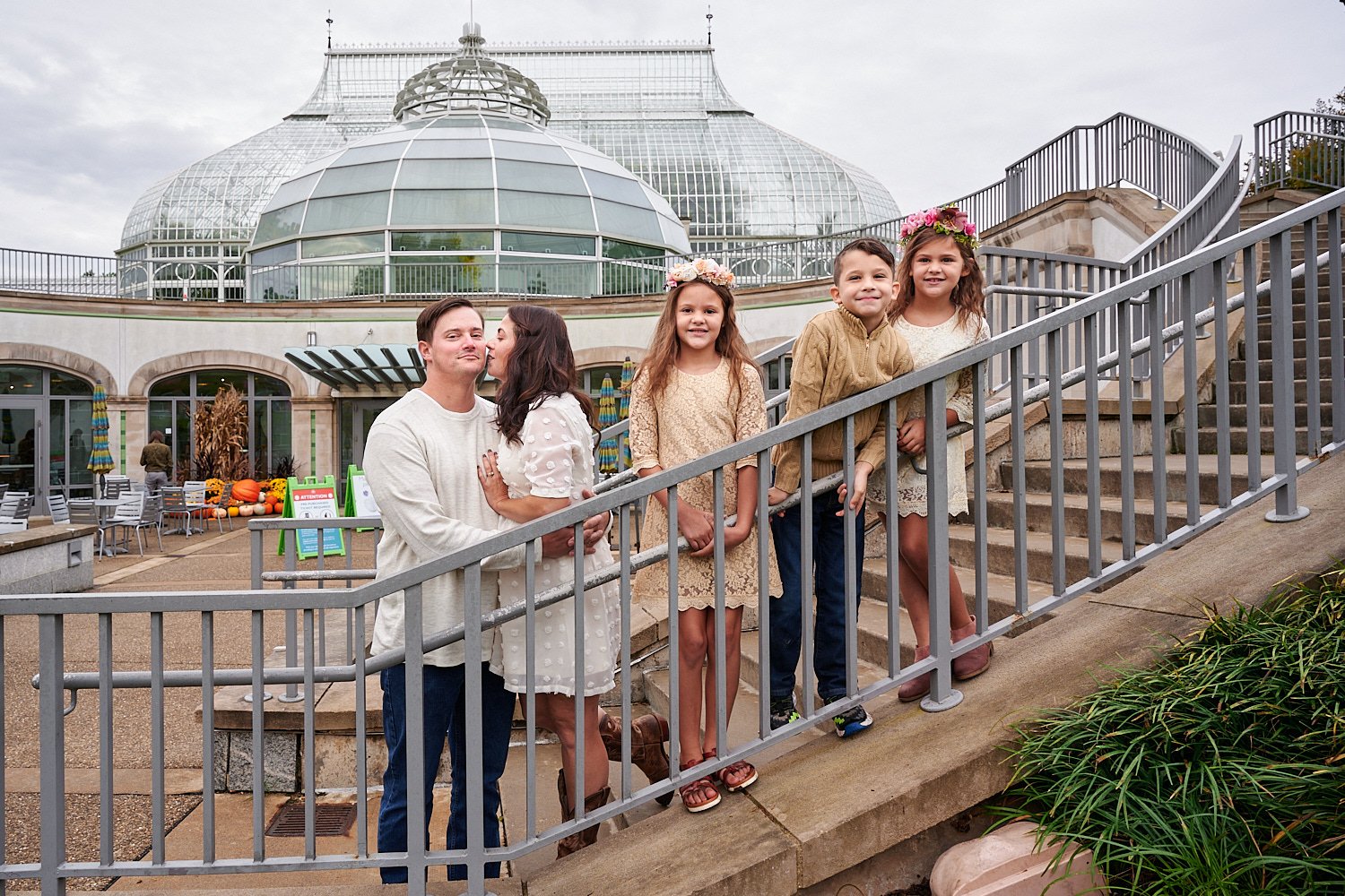 Zack Reed is posing with his wife, son and two daughters in front of Phipps Conservatory and Botanical Gardens of Pittsburgh, Pennsylvania. The ladies are wearing embroidered beige dresses and flowers 