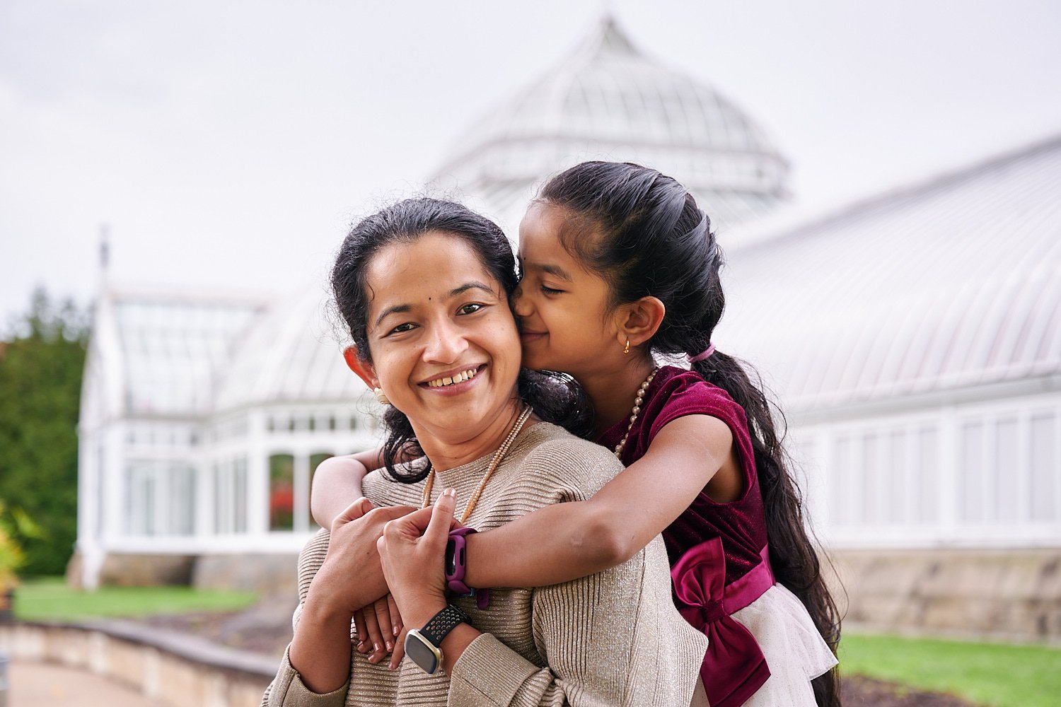  mother is posing with her pre-teen daughter in front of Phipps Conservatory and Botanical Gardens of Pittsburgh, Pennsylvania. The girl is wearing a burgundy ball dress and pearls. 