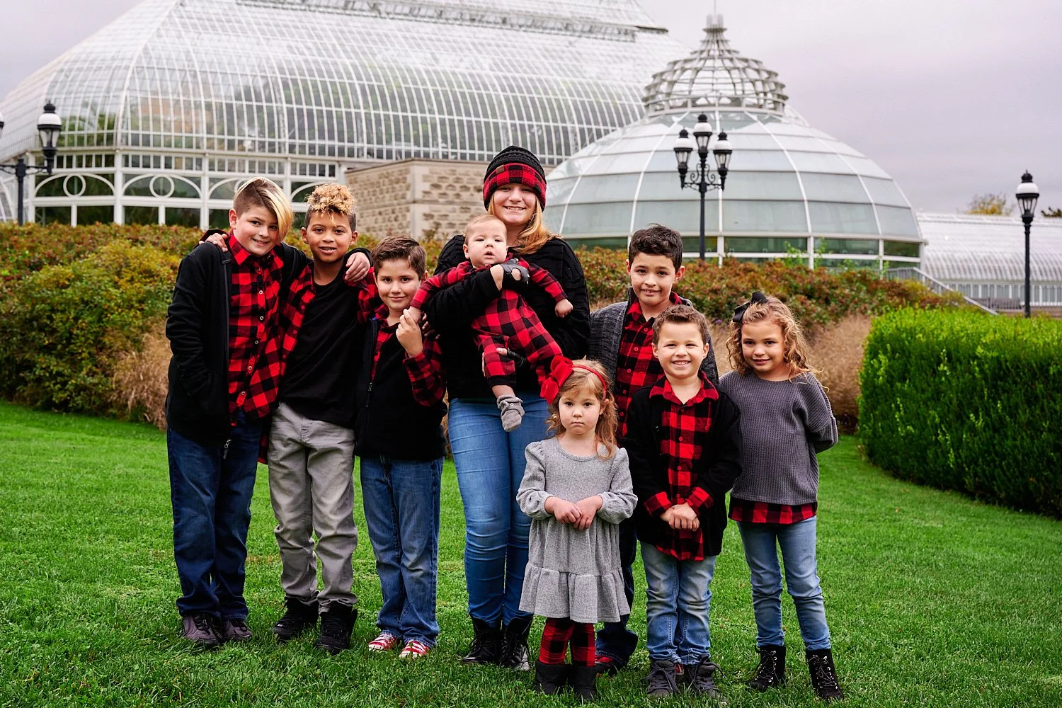  Andrea D’Alessandro is posing with her extended family in front of Phipps Conservatory and Botanical Gardens of Pittsburgh, PA. The large group is wearing red checked flannel shirts and blue jeans. 