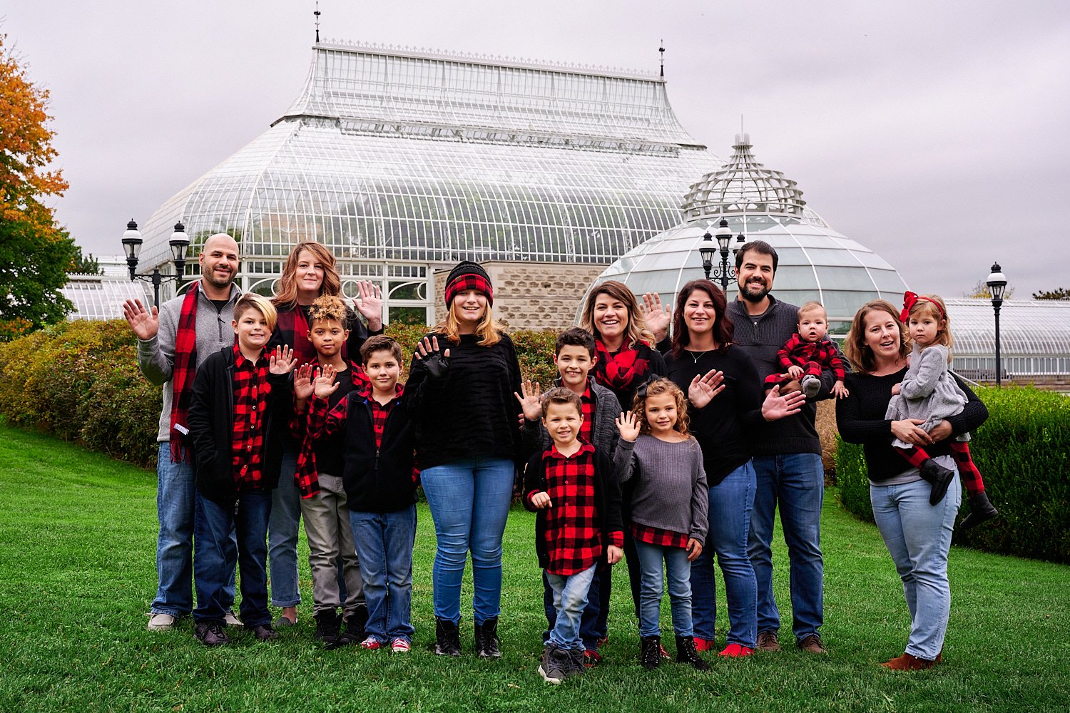 Andrea D’Alessandro is posing with her extended family in front of Phipps Conservatory and Botanical Gardens of Pittsburgh, PA. The large group is wearing red checked flannel shirts and blue jeans. 