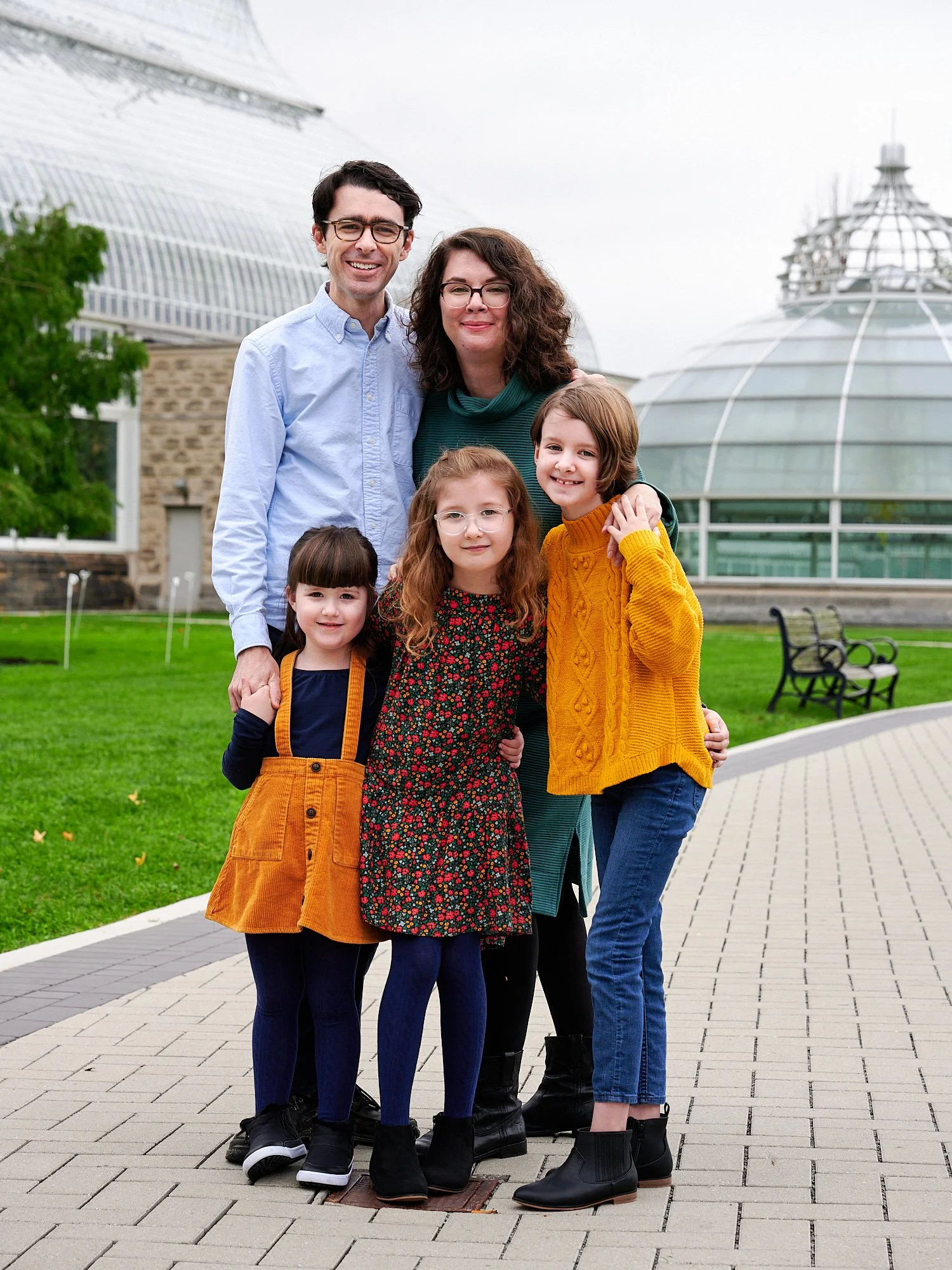  Spencer Kingman is posing with family in front of Phipps Conservatory and Botanical Gardens of Pittsburgh, PA. Mother and three daughters are wearing dresses in jewel colors - yellow, blue and green. 