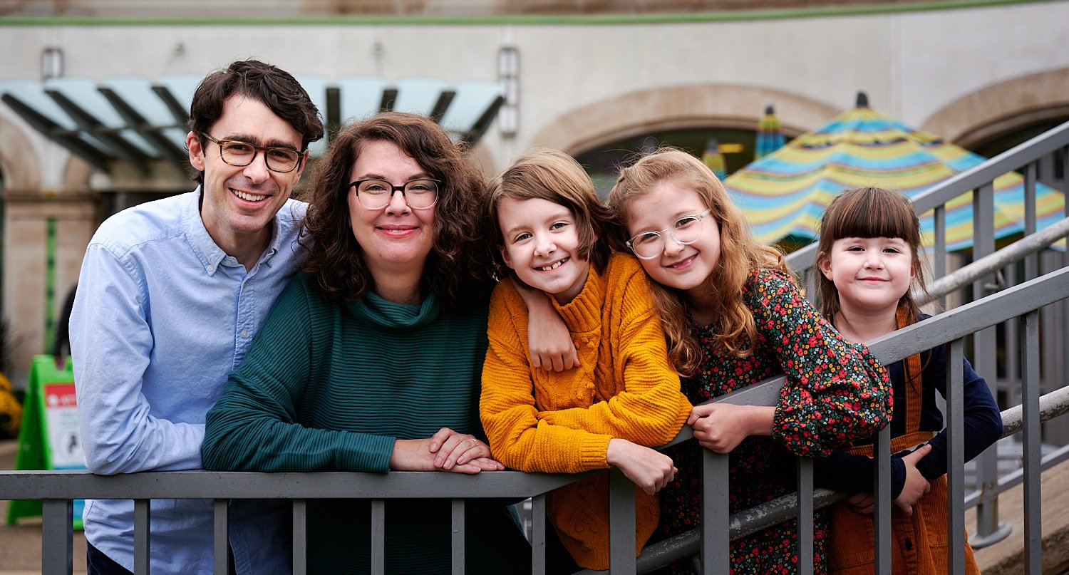  Spencer Kingman is posing with family in front of Phipps Conservatory and Botanical Gardens of Pittsburgh, PA. Mother and three daughters are wearing dresses in jewel colors - yellow, blue and green. 