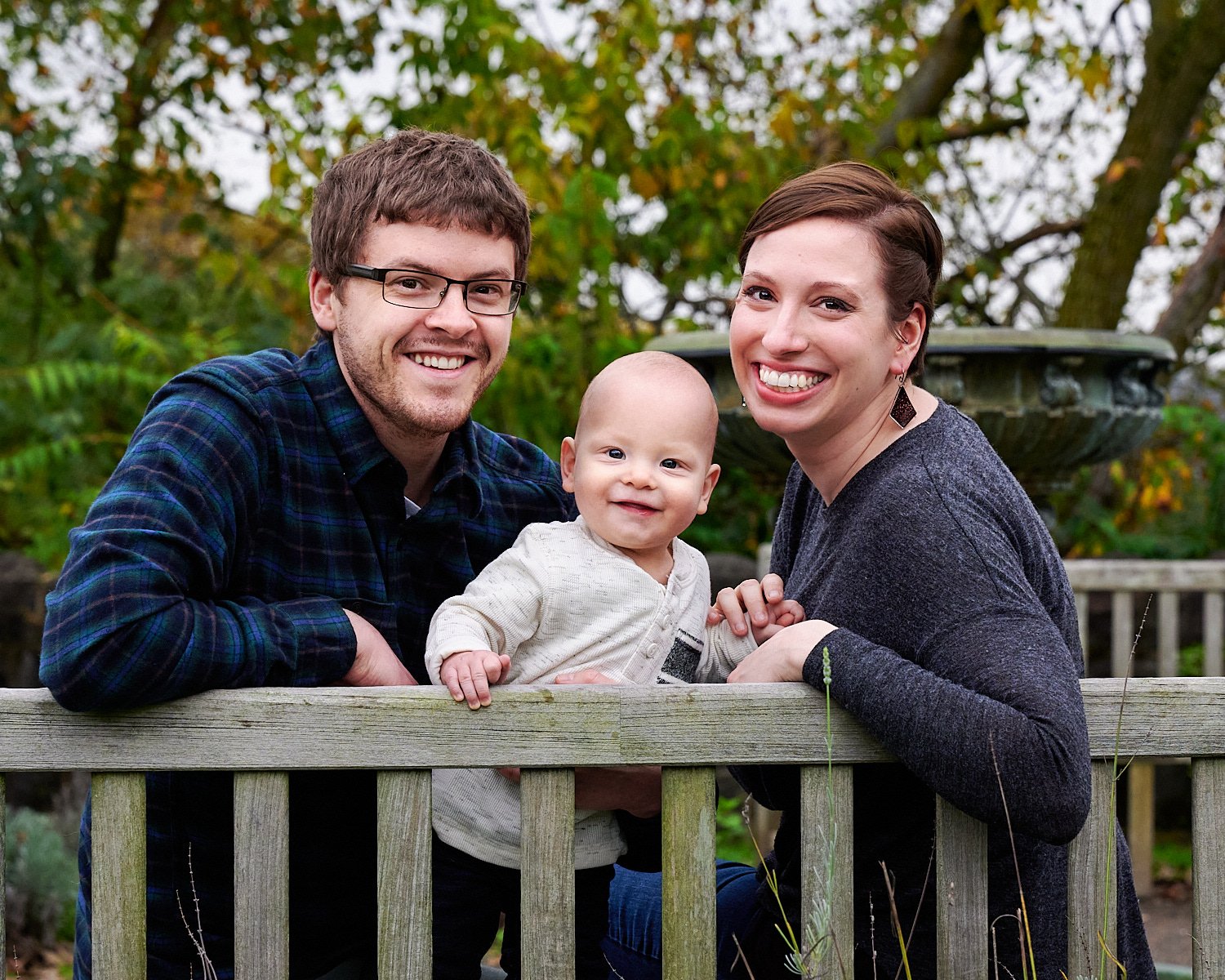  Leanne Elliott is posing with her bespectacled husband and a smiling 9 month old son in front of Phipps Conservatory and Botanical Gardens of Pittsburgh, PA. The family is dressed in a checked shirt. 