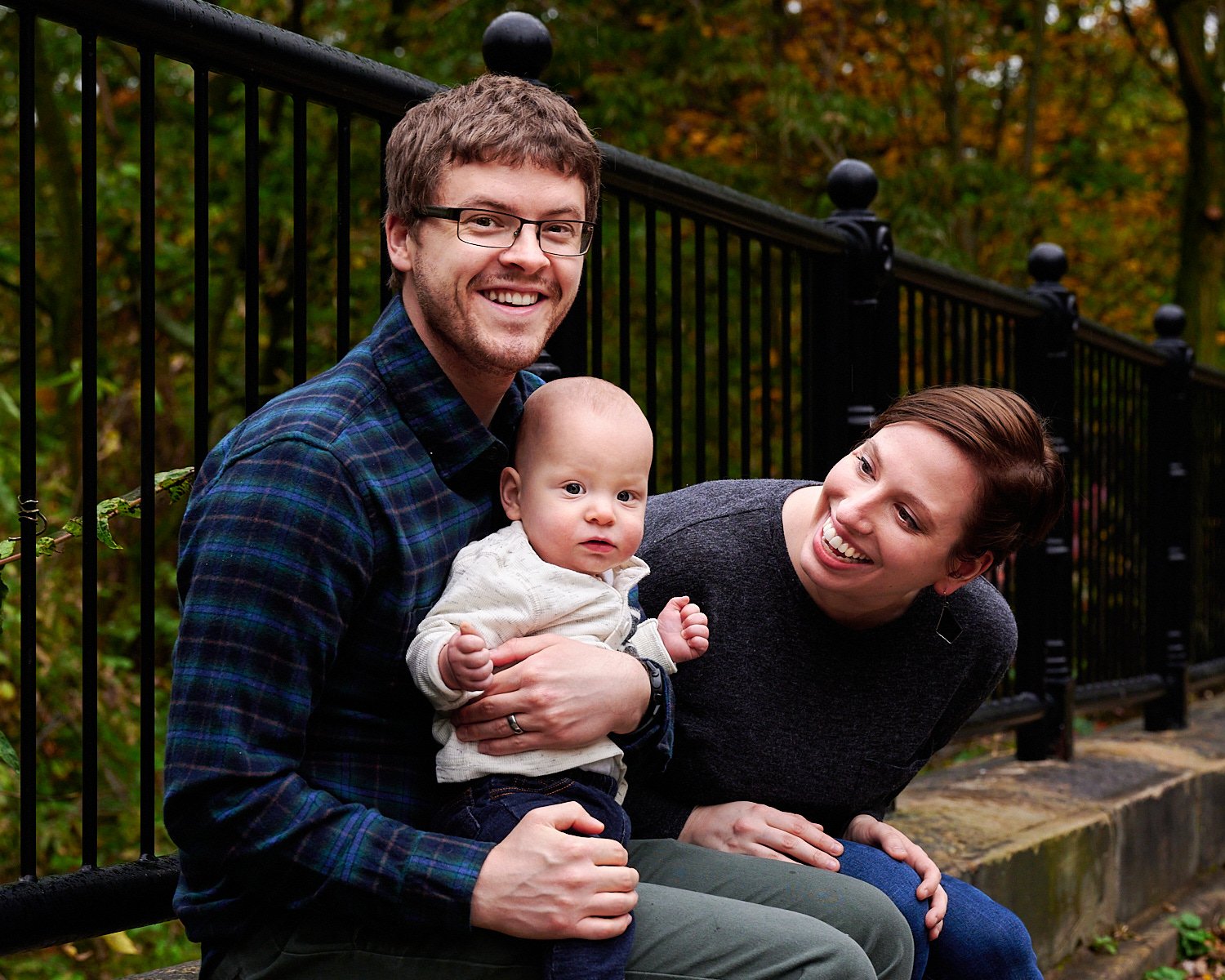  Leanne Elliott is posing with her bespectacled husband and a smiling 9 month old son in front of Phipps Conservatory and Botanical Gardens of Pittsburgh, PA. The family is dressed in a checked shirt. 