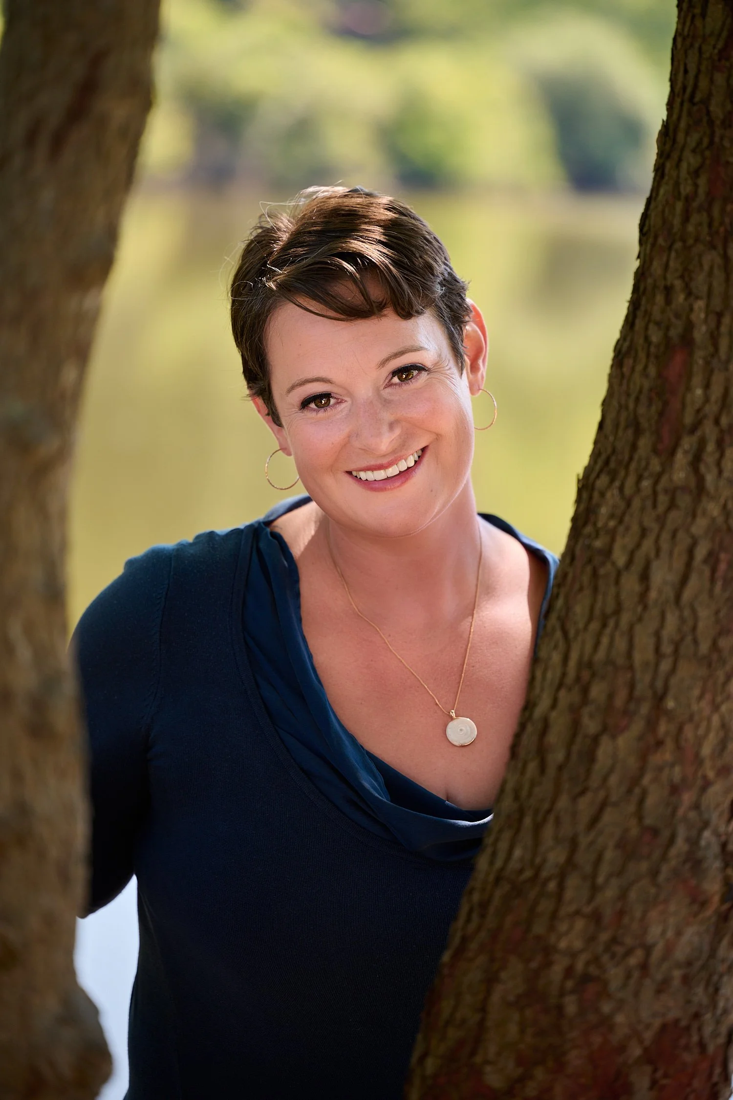 Rachael Zito is posing for business headshots and personal branding portraits on a warm sunny day in the park. She is smiling wide and standing in the tall grass and by a tree wearing a v-neck blouse 