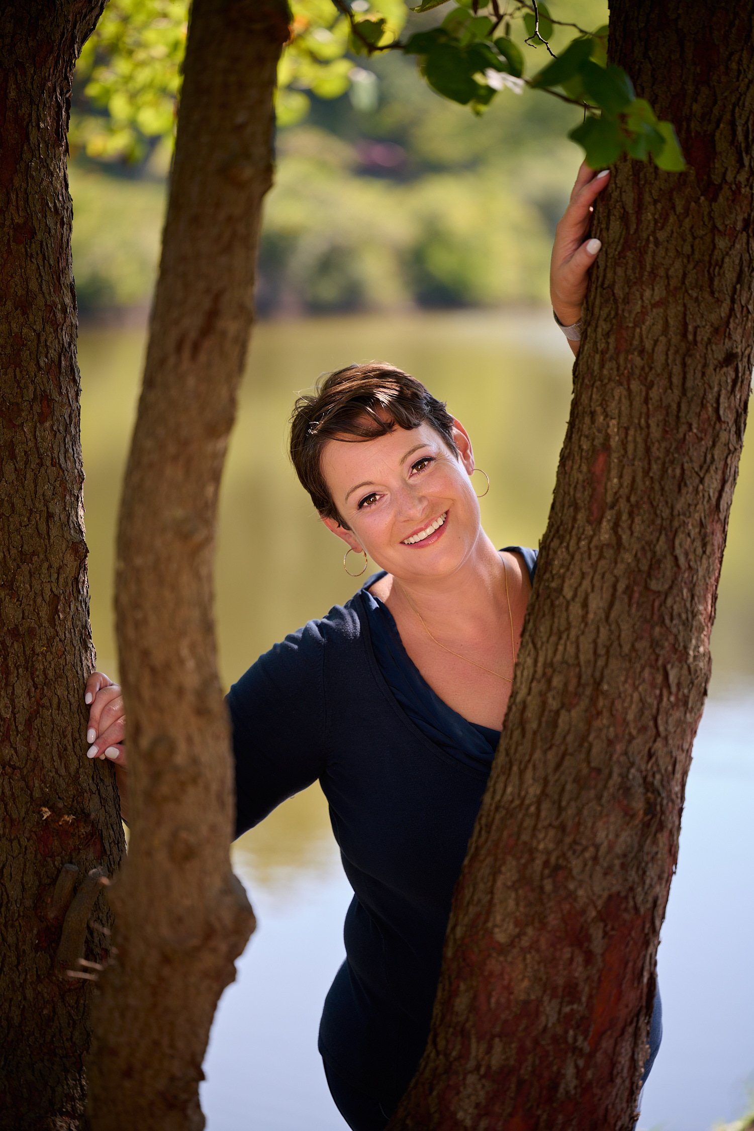  Rachael Zito is posing for business headshots and personal branding portraits on a warm sunny day in the park. She is smiling wide and standing in the tall grass and by a tree wearing a v-neck blouse 