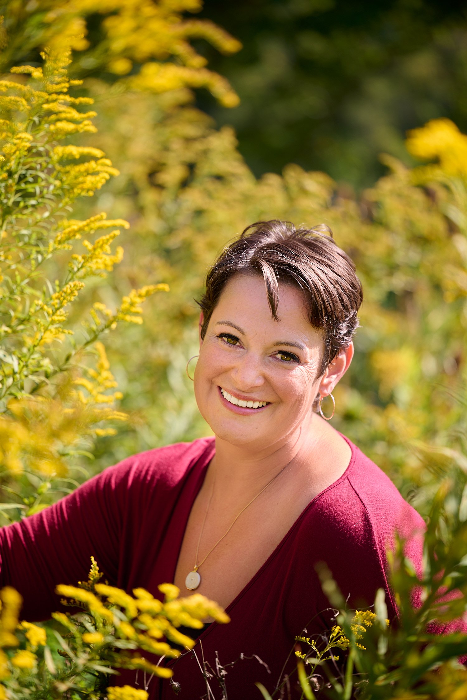  Rachael Zito is posing for business headshots and personal branding portraits on a warm sunny day in the park. She is smiling wide and standing in the tall grass and by a tree wearing a v-neck blouse 