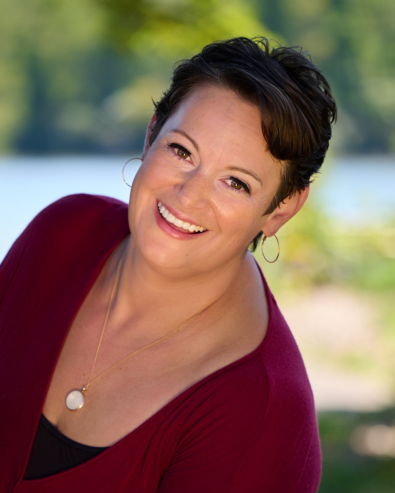  Rachael Zito is posing for business headshots and personal branding portraits on a warm sunny day in the park. She is smiling wide and standing in the tall grass and by a tree wearing a v-neck blouse 