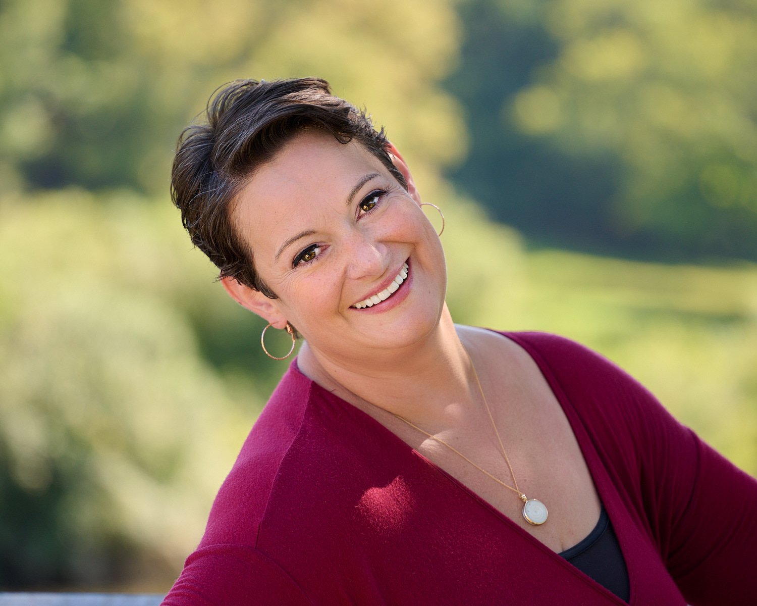  Rachael Zito is posing for business headshots and personal branding portraits on a warm sunny day in the park. She is smiling wide and standing in the tall grass and by a tree wearing a v-neck blouse 