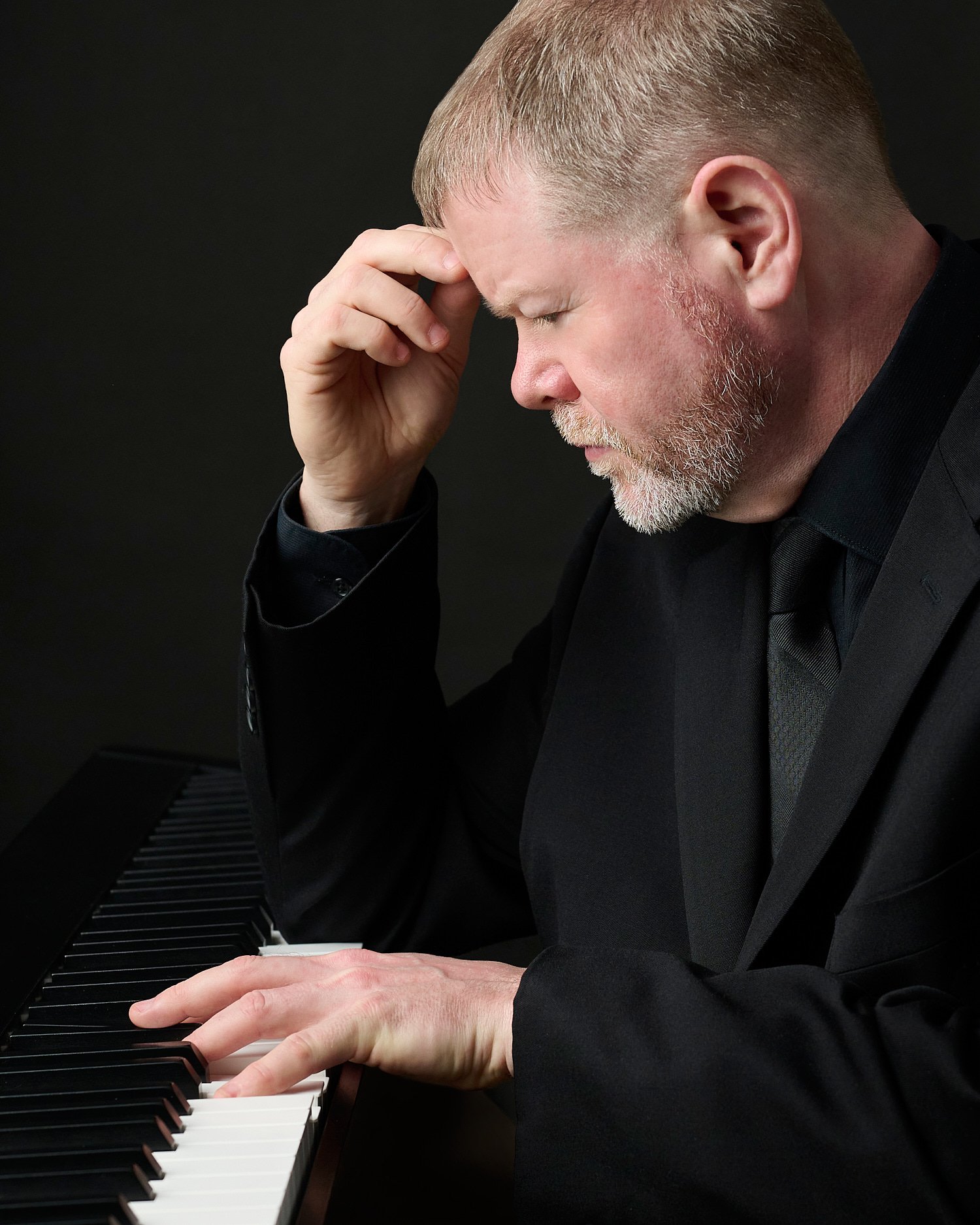 Tim Heavner is posing for his business headshots in a professional photography studio with a dark black background. Timothy is a pianist and is portrayed with his keyboard full length and close-up. 