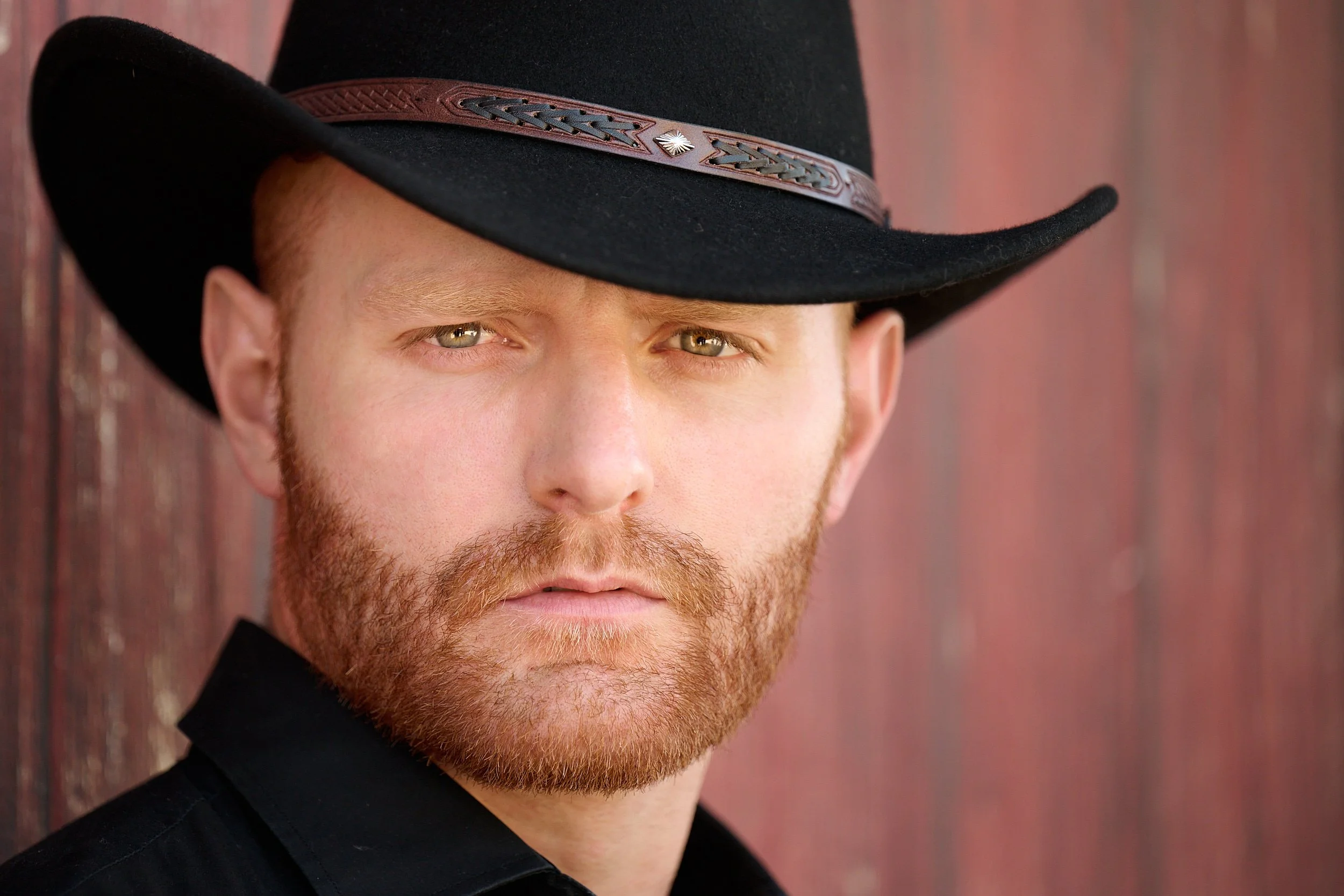 THE WOODLANDS, TEXAS - MAY 2022: Wyatt Hopkins is posing for sophisticated cowboy portraits by a barn on a  horse ranch. The man has short red hair, mustache and beard. He wears a black fedora hat. 