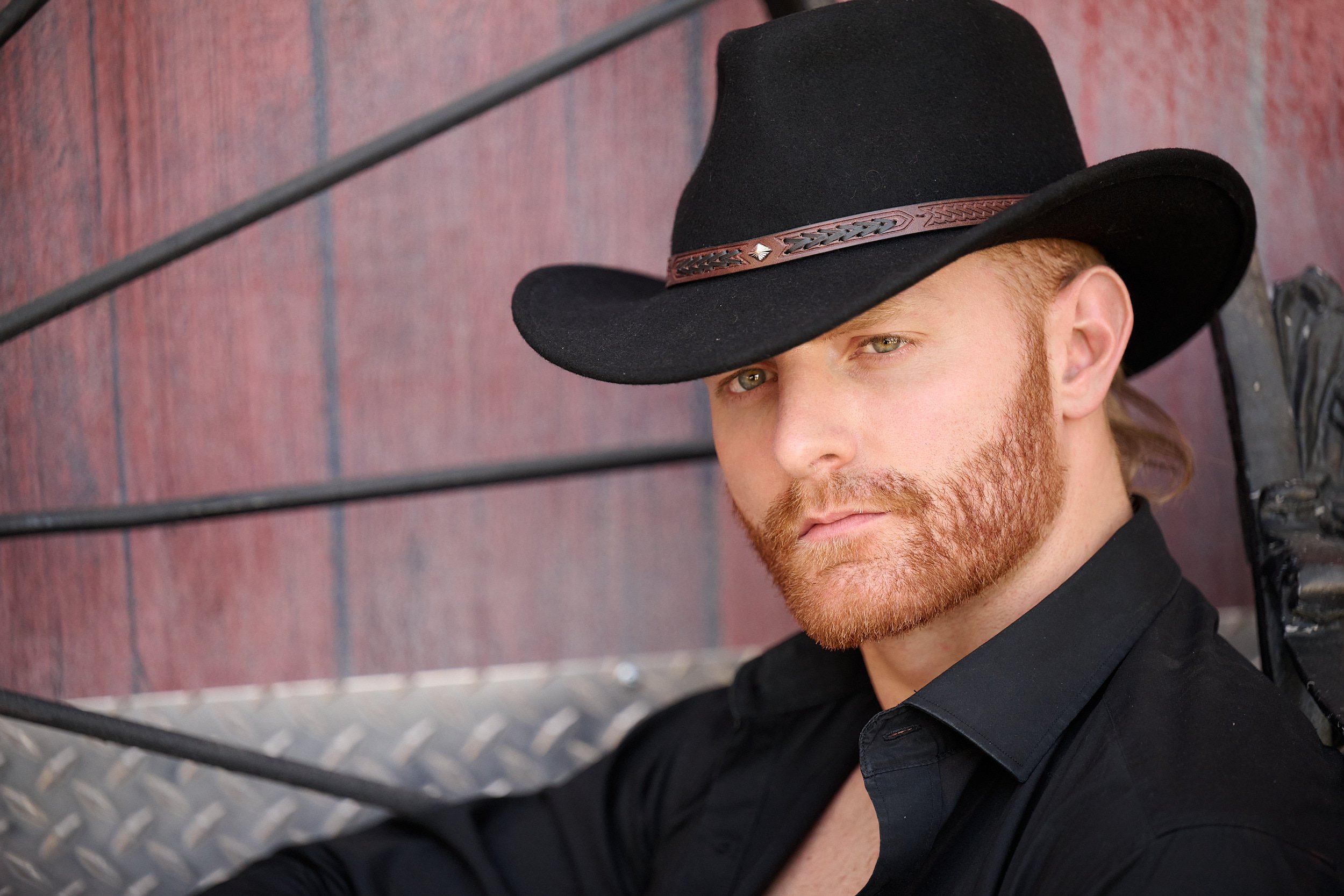  THE WOODLANDS, TEXAS - MAY 2022: Wyatt Hopkins is posing for sophisticated cowboy portraits by a barn on a  horse ranch. The man has short red hair, mustache and beard. He wears a black fedora hat. 