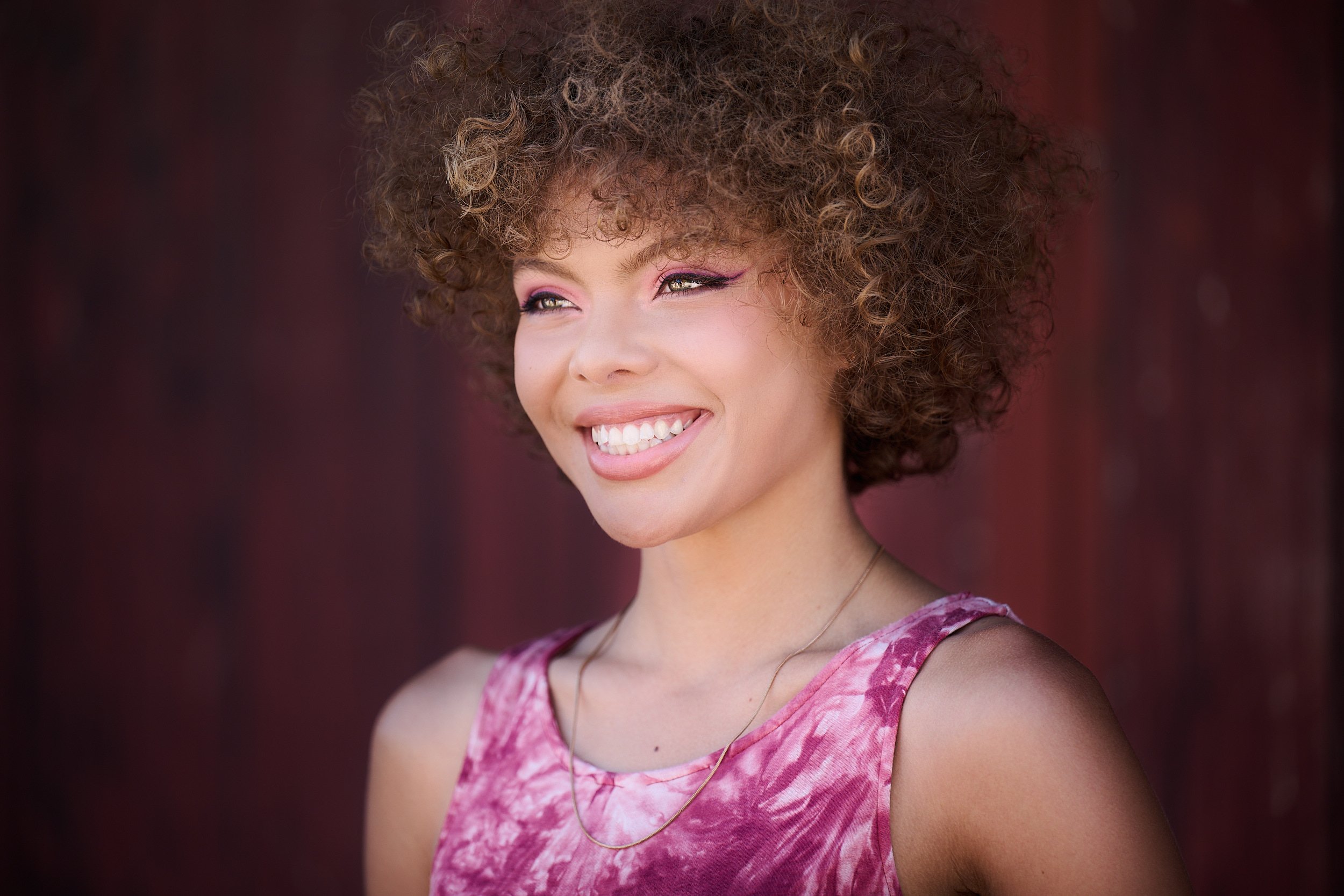  THE WOODLANDS, TEXAS - MAY 2022: Journee Reid is posing for formal and elegant portraits by a rustic wall. The young girl has short curly hair sunlit from above. She wears a pink sleeveless dress. 