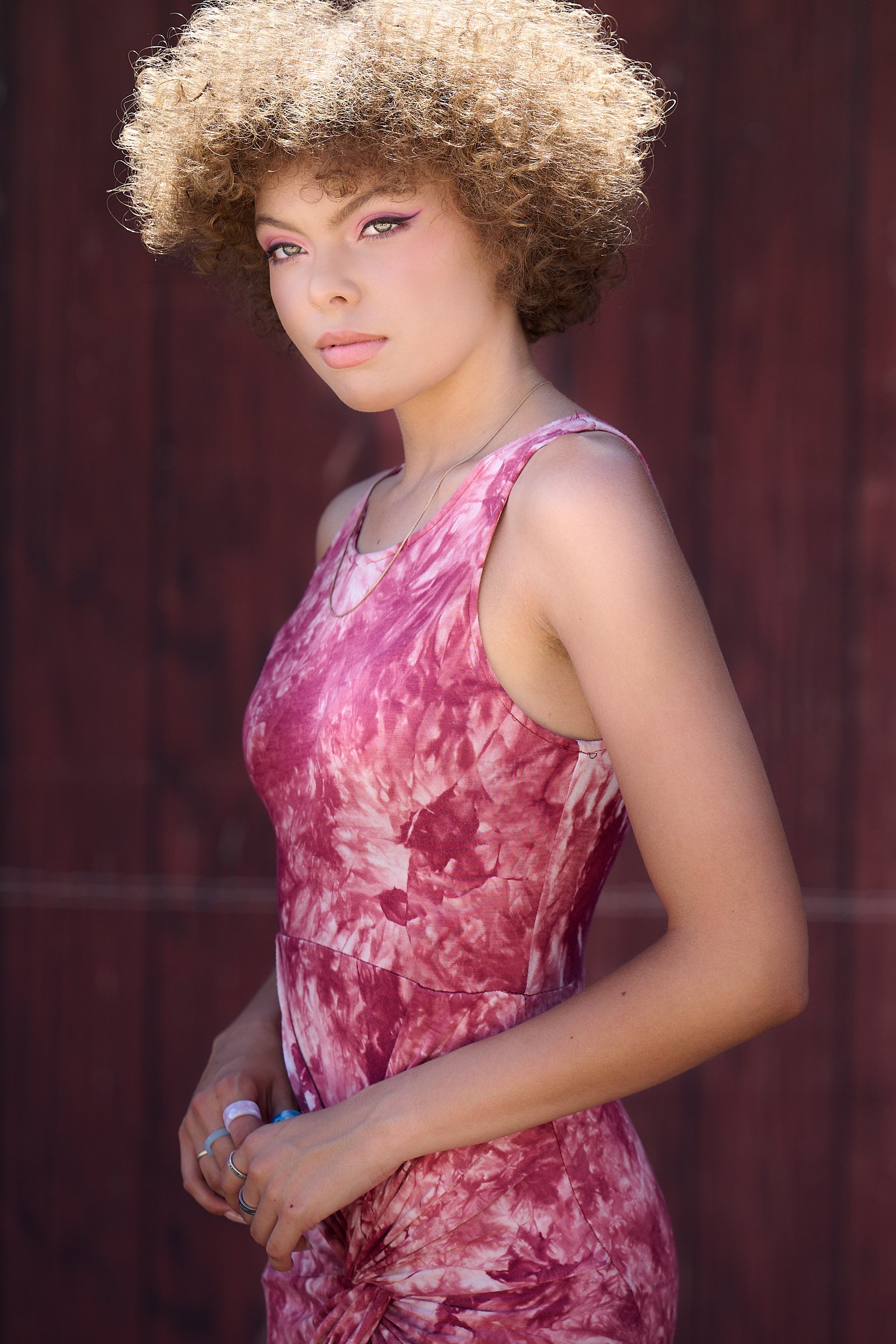  THE WOODLANDS, TEXAS - MAY 2022: Journee Reid is posing for formal and elegant portraits by a rustic wall. The young girl has short curly hair sunlit from above. She wears a pink sleeveless dress. 