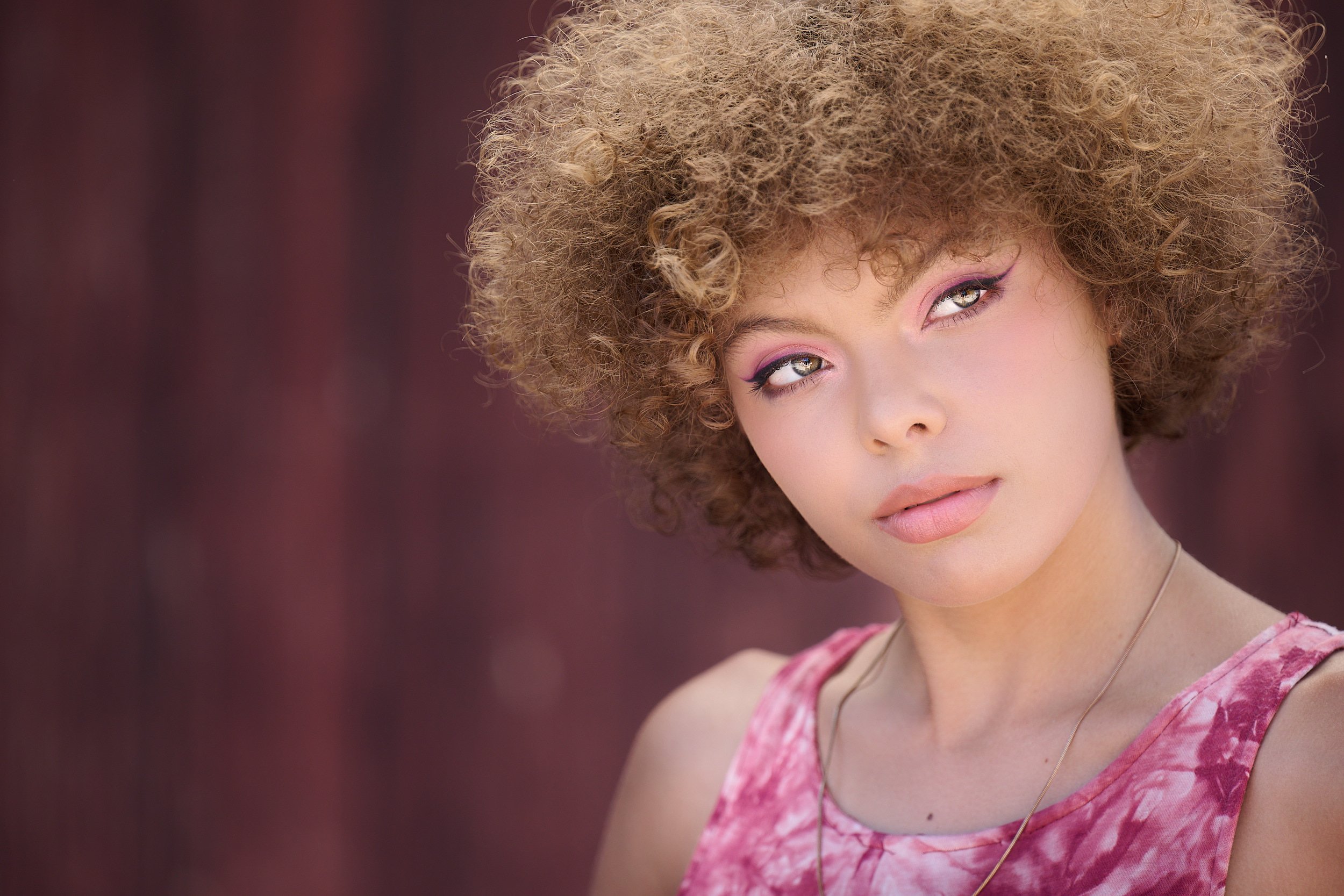  THE WOODLANDS, TEXAS - MAY 2022: Journee Reid is posing for formal and elegant portraits by a rustic wall. The young girl has short curly hair sunlit from above. She wears a pink sleeveless dress. 