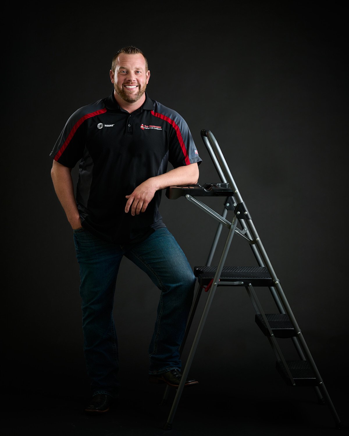  THE WOODLANDS, TEXAS - May 16 2023: Austin Jones of The Woodlands Heating and Air Conditioning is posing against a black background by a ladder dressed in jeans and a polo shirt with his company logo 