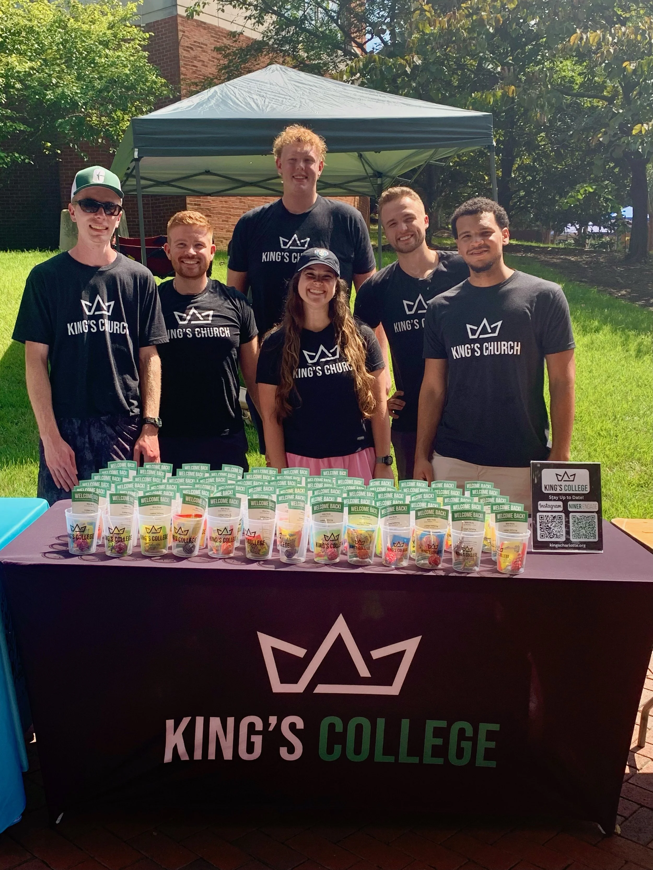 Several smiling young adults standing behind a table with cups featuring King's College logo and slogans, under a canopy, outdoors on a sunny day, with trees and a brick building in the background.