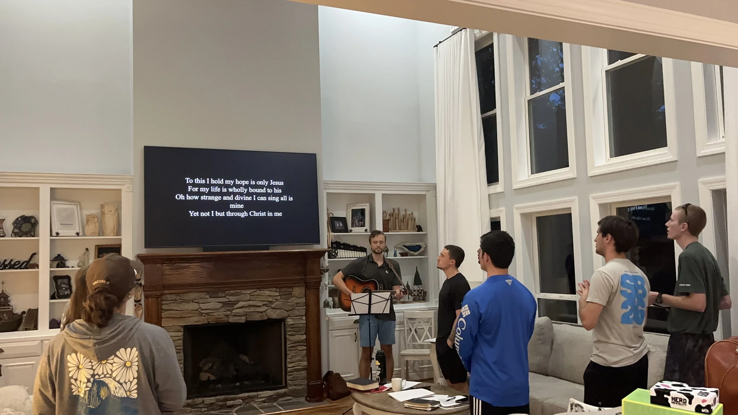 A group of people standing in a living room singing and playing guitar during a gathering, with a large screen displaying song lyrics on the wall.