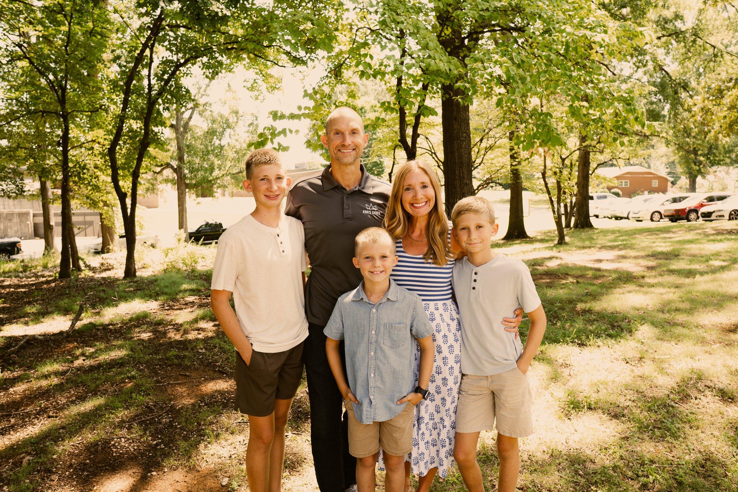 A smiling family of six standing outdoors under a canopy of trees on a sunny day, with parked cars in the background.