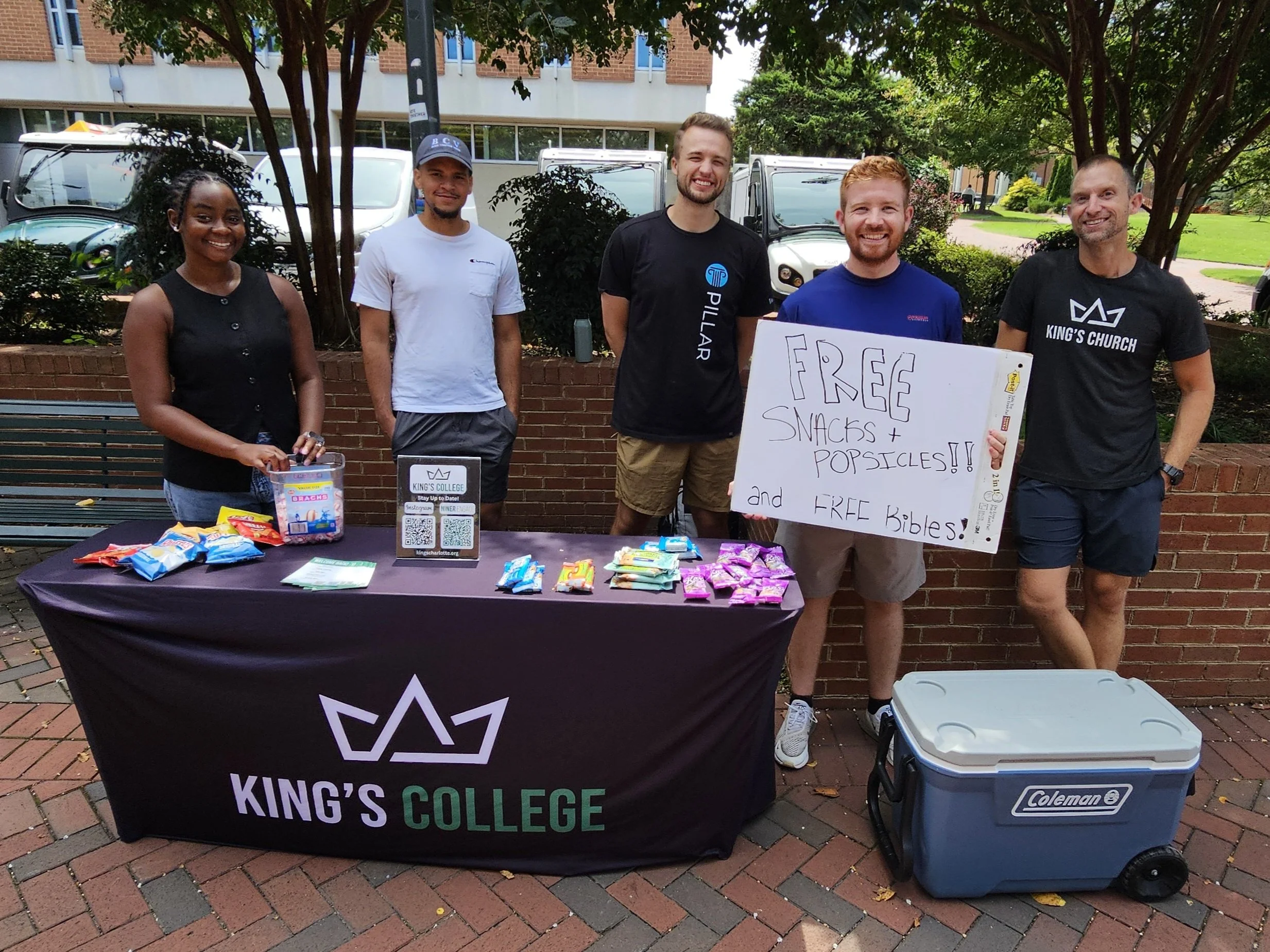 A group of five people standing behind a table with free snacks and popsicles at UNCC. They are outdoors, under trees, with cars and bushes in the background. One person holds a whiteboard sign advertising free snacks, popsicles, and Bibles.