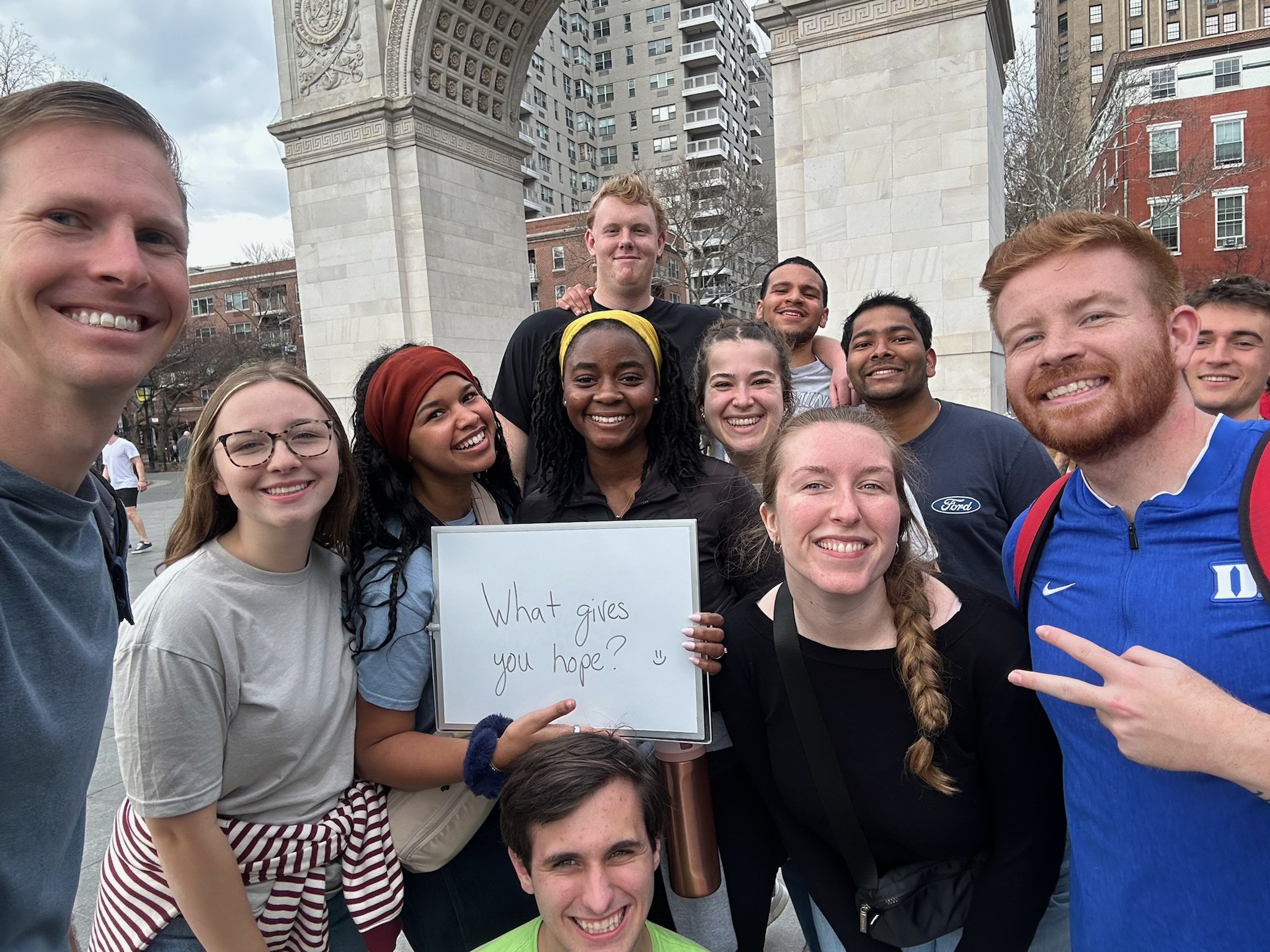 A group of young adults smiling and taking a selfie in a city park, with a historical arch monument and modern high-rise buildings in the background. One person is holding a whiteboard with a handwritten question, 'What gives you hope?'