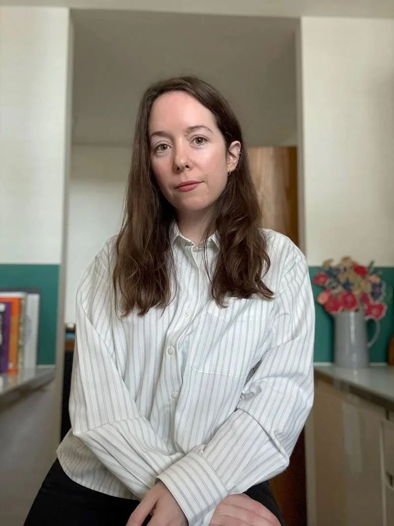 A woman with long brown hair and a neutral expression, wearing a white striped button-up shirt, sitting in a room with a bookshelf, a door, and a vase of colorful flowers in the background.