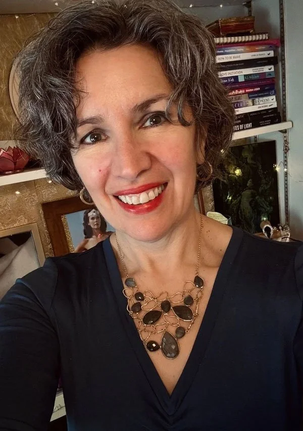 A woman with short curly gray hair smiling at the camera, wearing a dark top and a statement necklace with black stones, in a room with books and decorative items in the background.