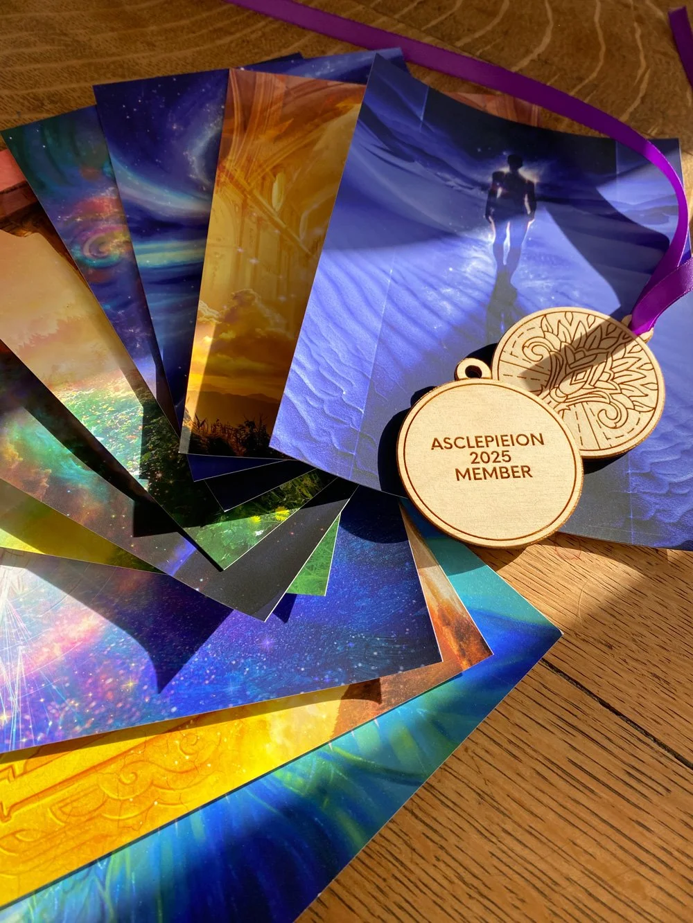 A collection of colorful inspirational cards with cosmic and nature themes, along with two wooden tokens labeled "ASCLIPLEION 2025 MEMBER."