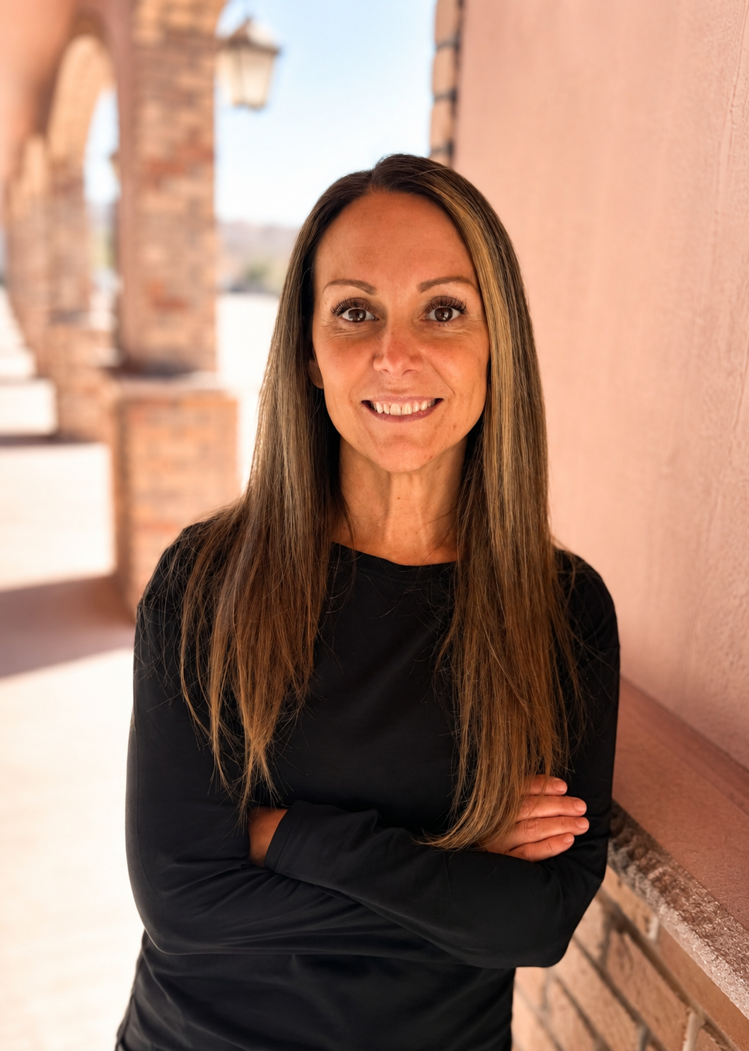 A woman with long brown hair, wearing a black long-sleeve top, smiling with her arms crossed, standing against a brick wall in a sunny outdoor setting.