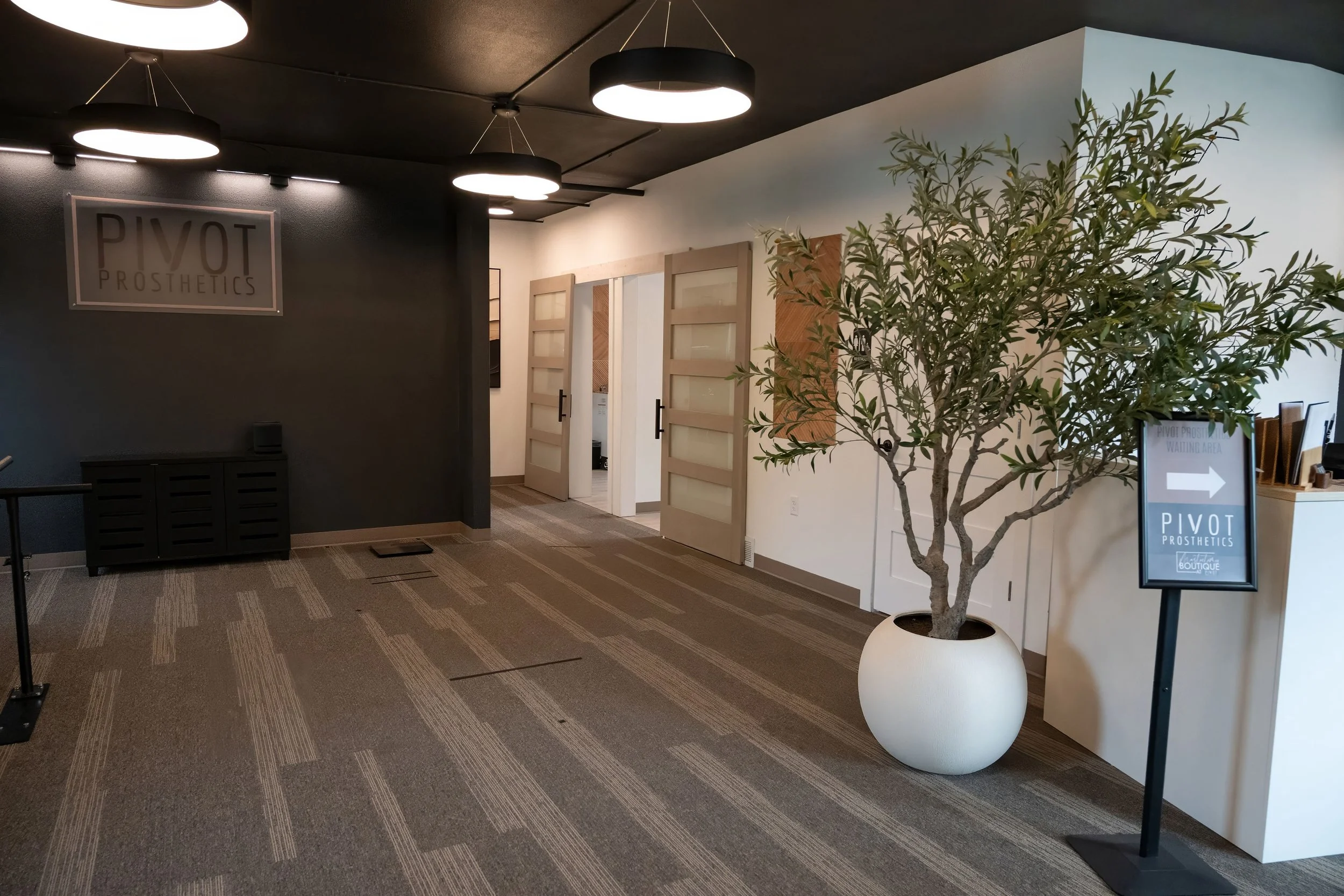 Interior of a modern office or clinic with a black ceiling and dark gray walls, illuminated by circular ceiling lights. There is a sign on the wall reading 'PIVOT PROSTHETICS.' A potted tree is in the foreground, and a sign with an arrow points to the right indicating the waiting area for PIVOT PROSTHETICS.