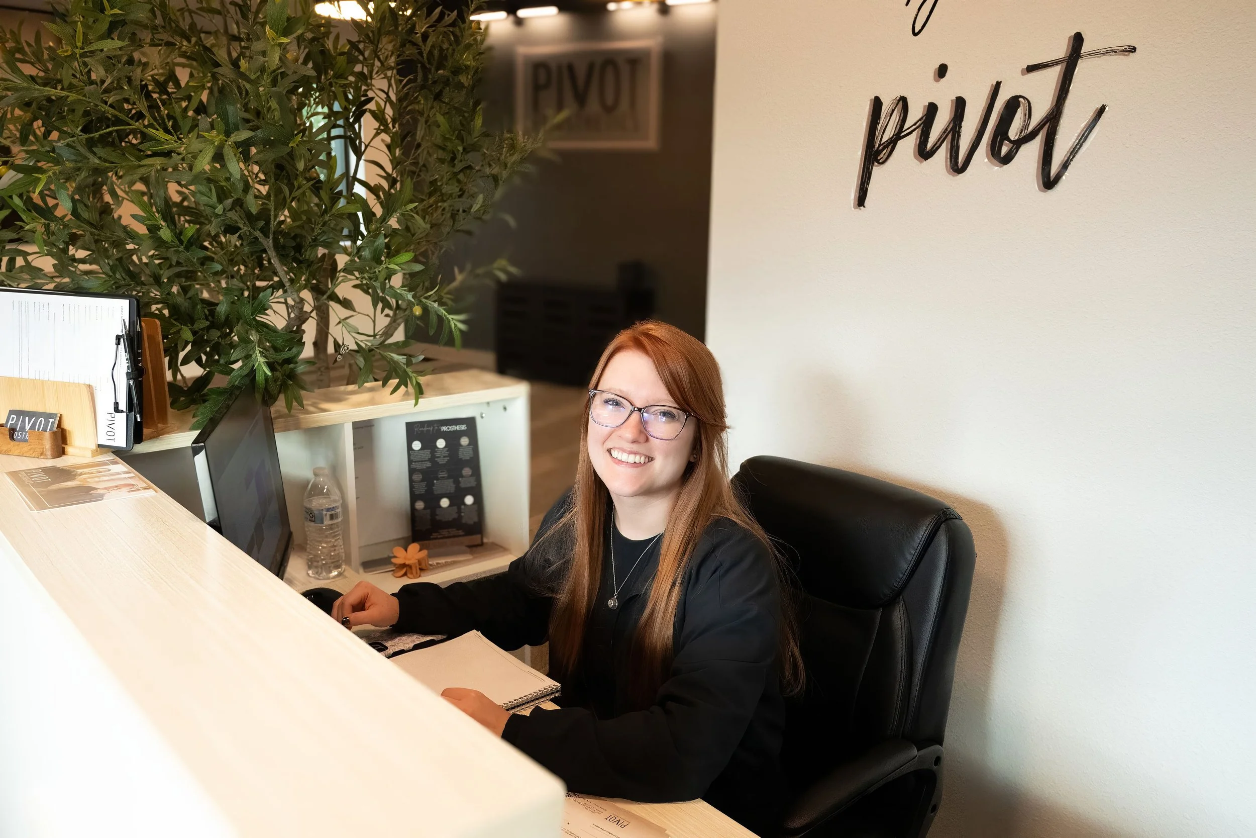 A young woman with red hair and glasses smiling while sitting at a reception desk in a modern office or cafe. Behind her is a large green plant, and on the wall to her right is a decorative sign that reads 'pivo' in cursive black letters. The desk has a computer monitor, a water bottle, and a small plant. There is also a menu or informational display on the counter.