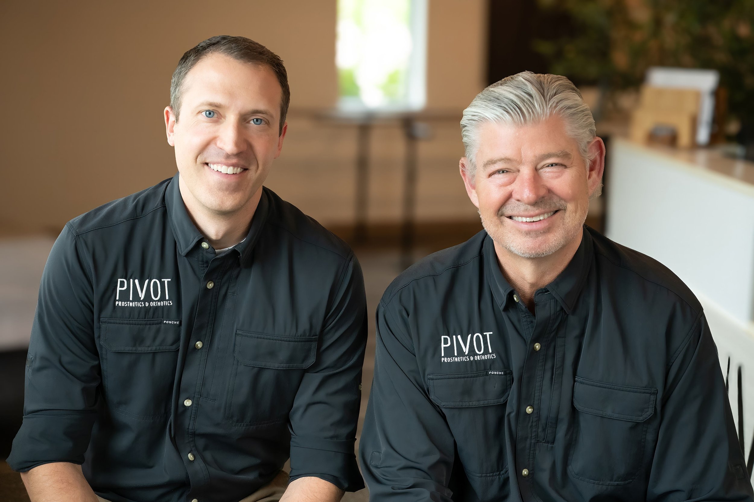 Brodie and Randy wearing black shirts with logo 'PIVOT Prosthetics & Orthotics' sitting indoors with a blurred background, ready to help provide a new leg or new arm after amputation in Rapid City, South Dakota