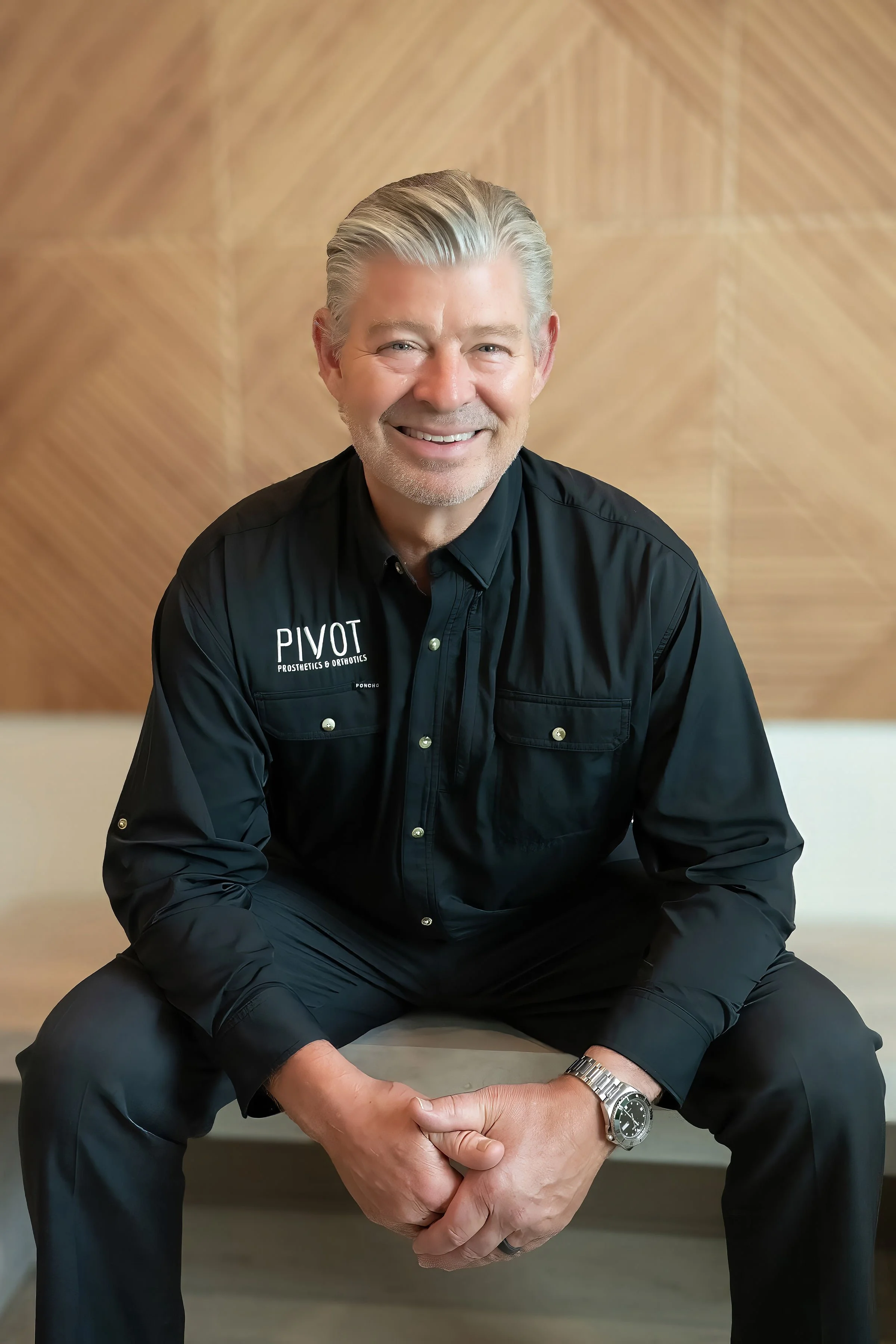 A smiling man with gray hair, wearing a black shirt with 'PIVOT Prosthetics & Orthotics' embroidered on it, sitting cross-legged against a wooden wall background.