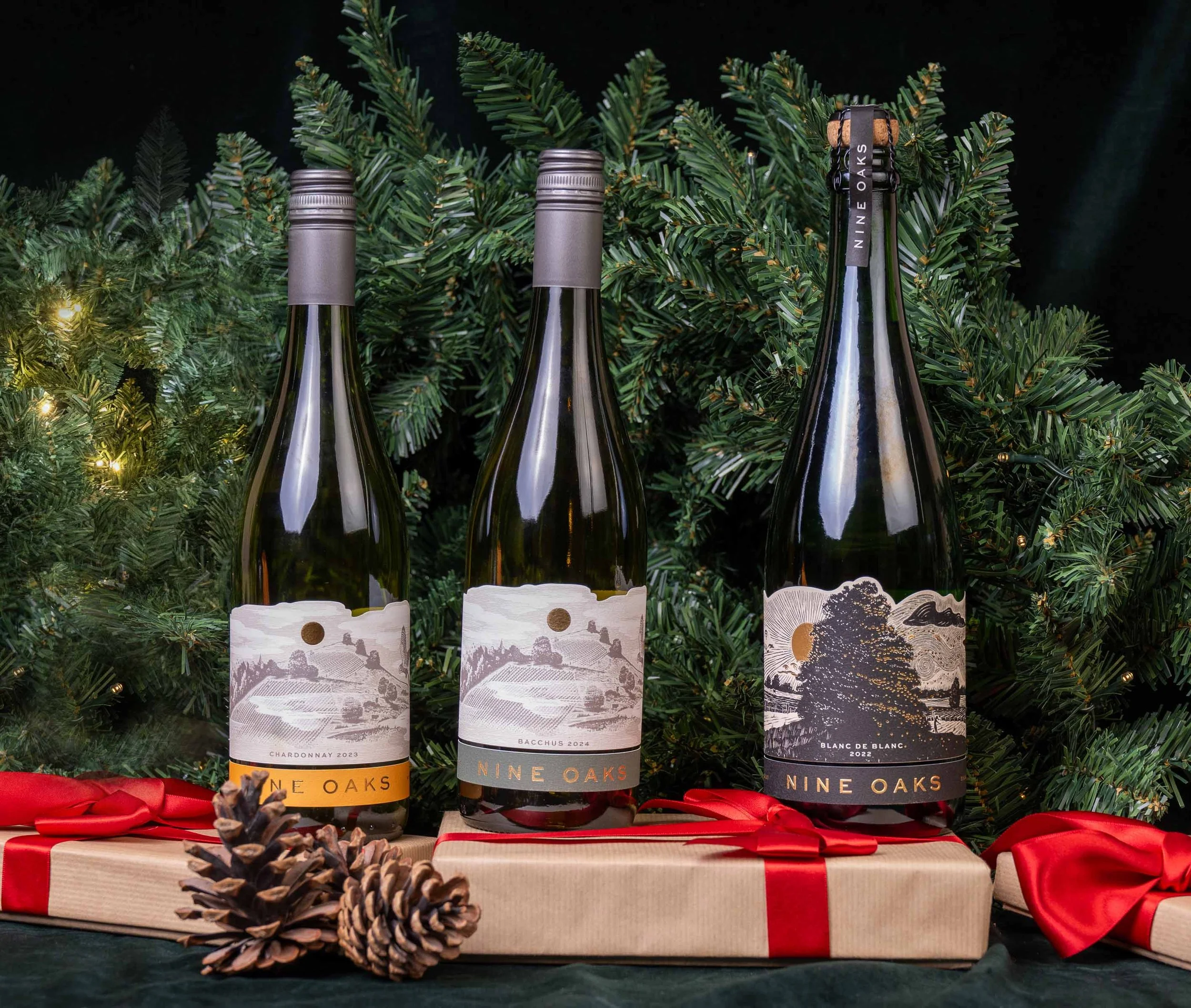 Three bottles of Nine Oaks wine (Chardonnay 2023, Bacchus 2024, Blanc de Blanc 2022) are displayed in front of green holiday Christmas tree branches with lights. The bottles are placed on a wrapped gift with a red ribbon, and pinecones and red ribbon bows are also present.