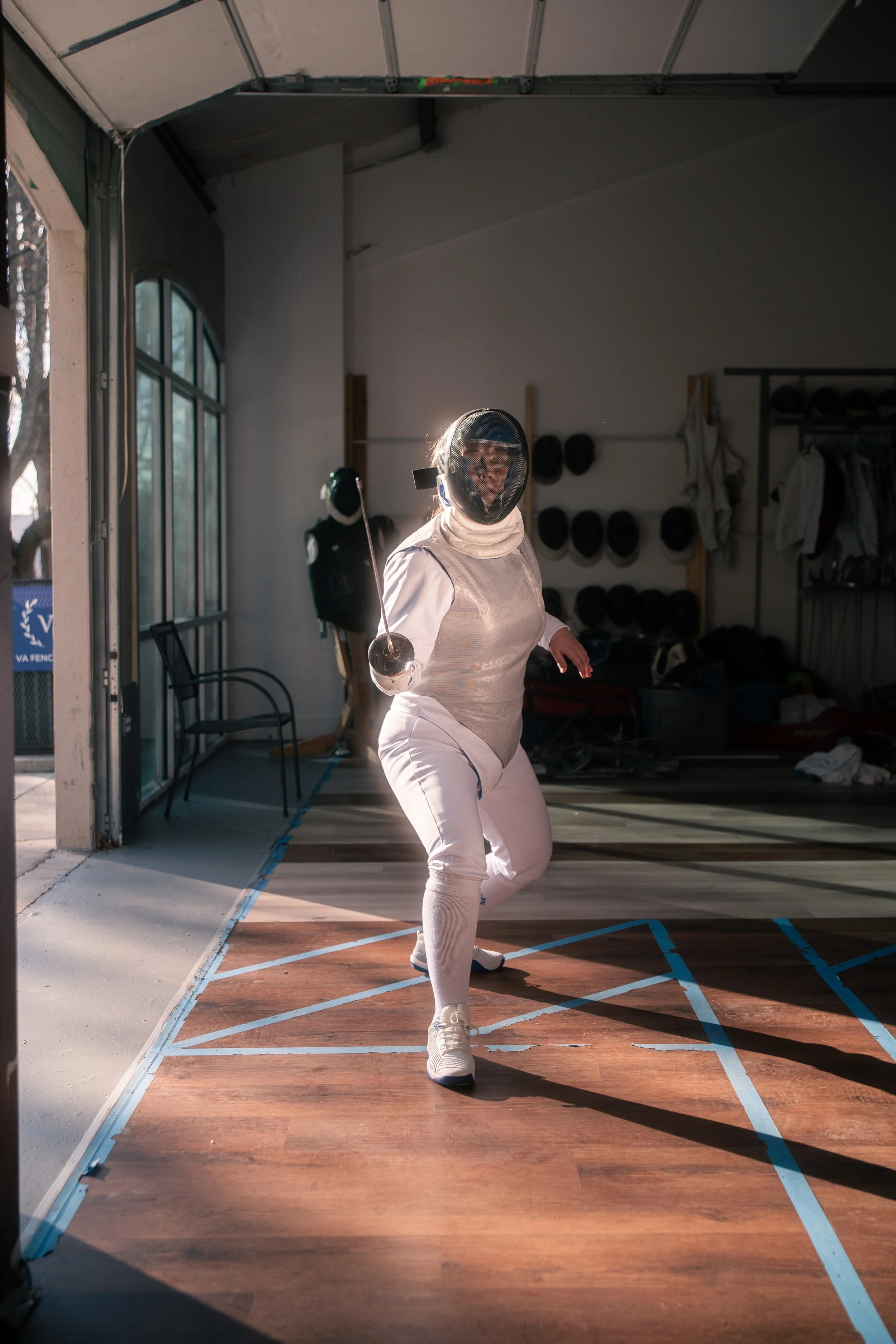 Female fencer practicing in indoor fencing gym, wearing protective gear, holding a foil.