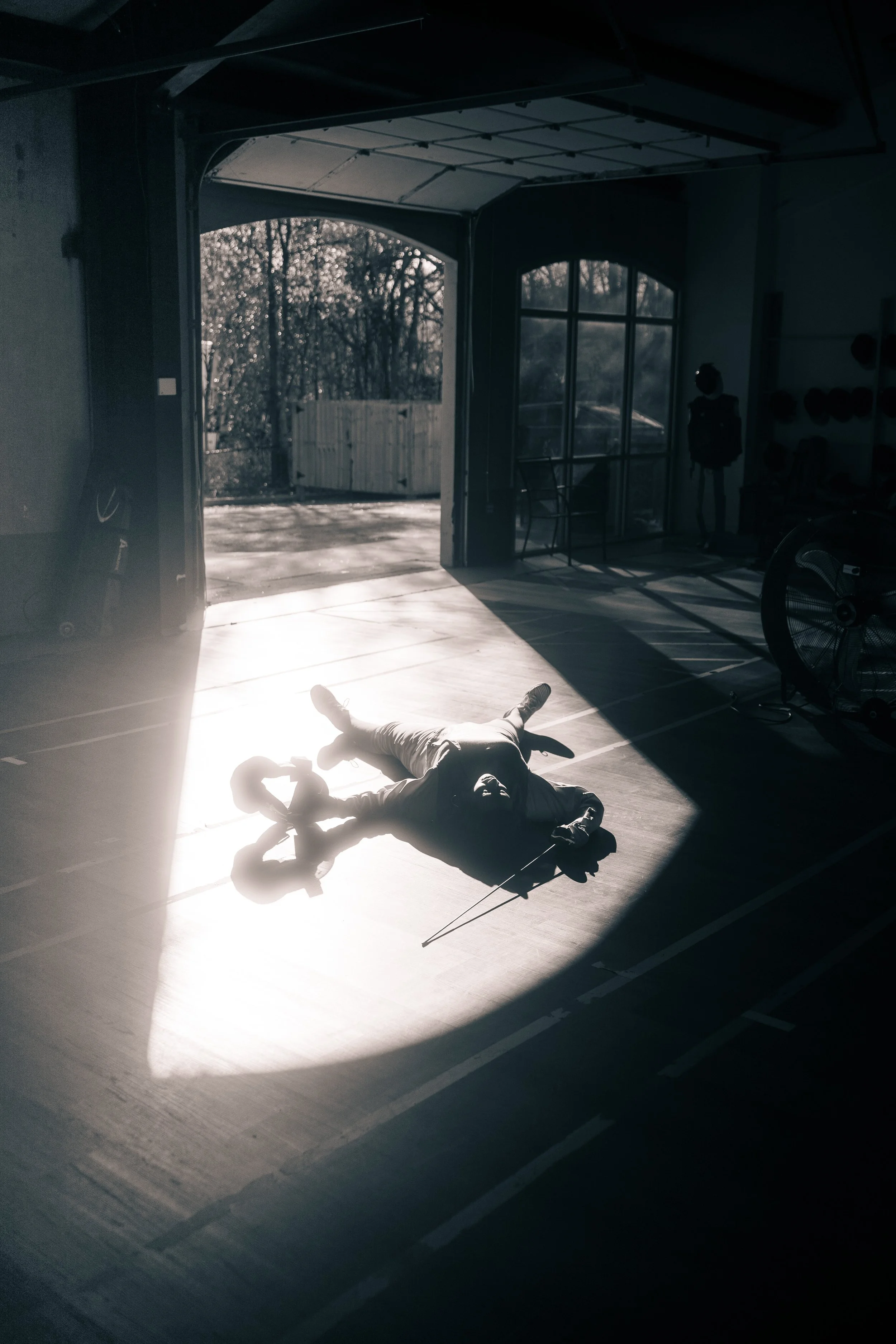 A person lying on the floor of a gym, illuminated by sunlight streaming through an open garage door, holding a fencing sword, with a mannequin and exercise equipment visible inside the gym.