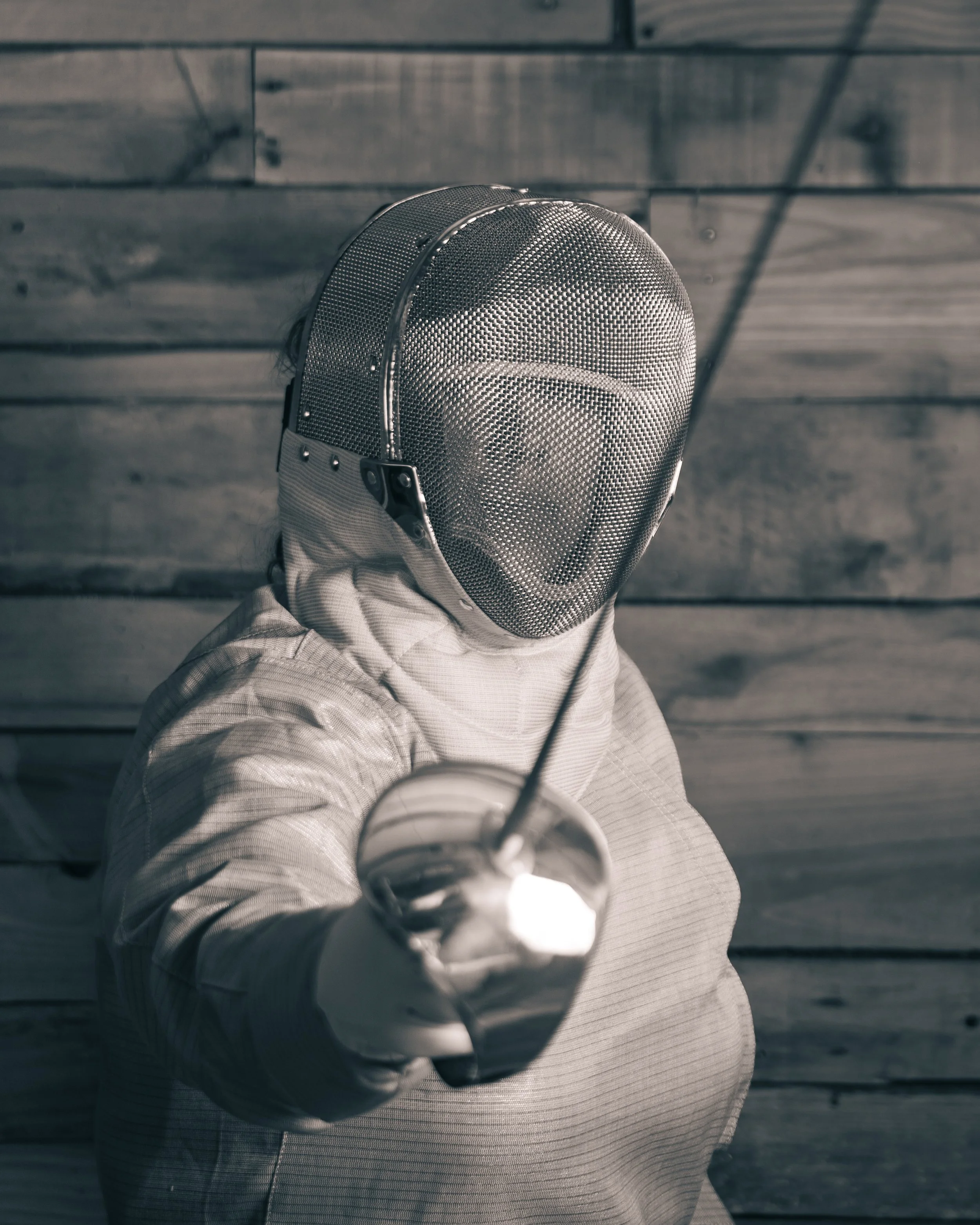 Person wearing fencing gear including a mask and jacket, holding a saber directed at the camera, with a wooden wall in the background.