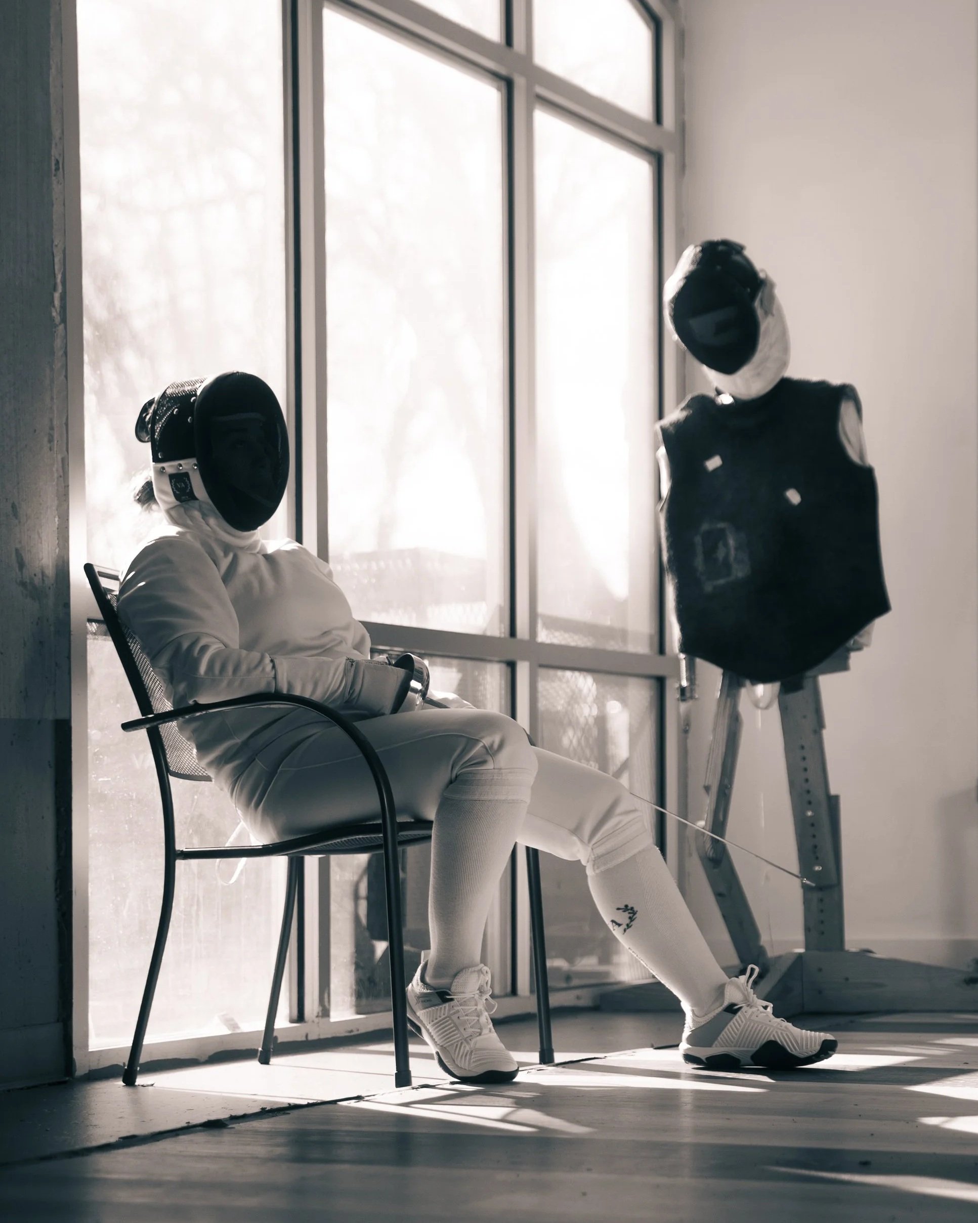 A person dressed in fencing gear, sitting on a chair, with a practice dummy nearby, positioned in front of large windows allowing natural light to fill the space.