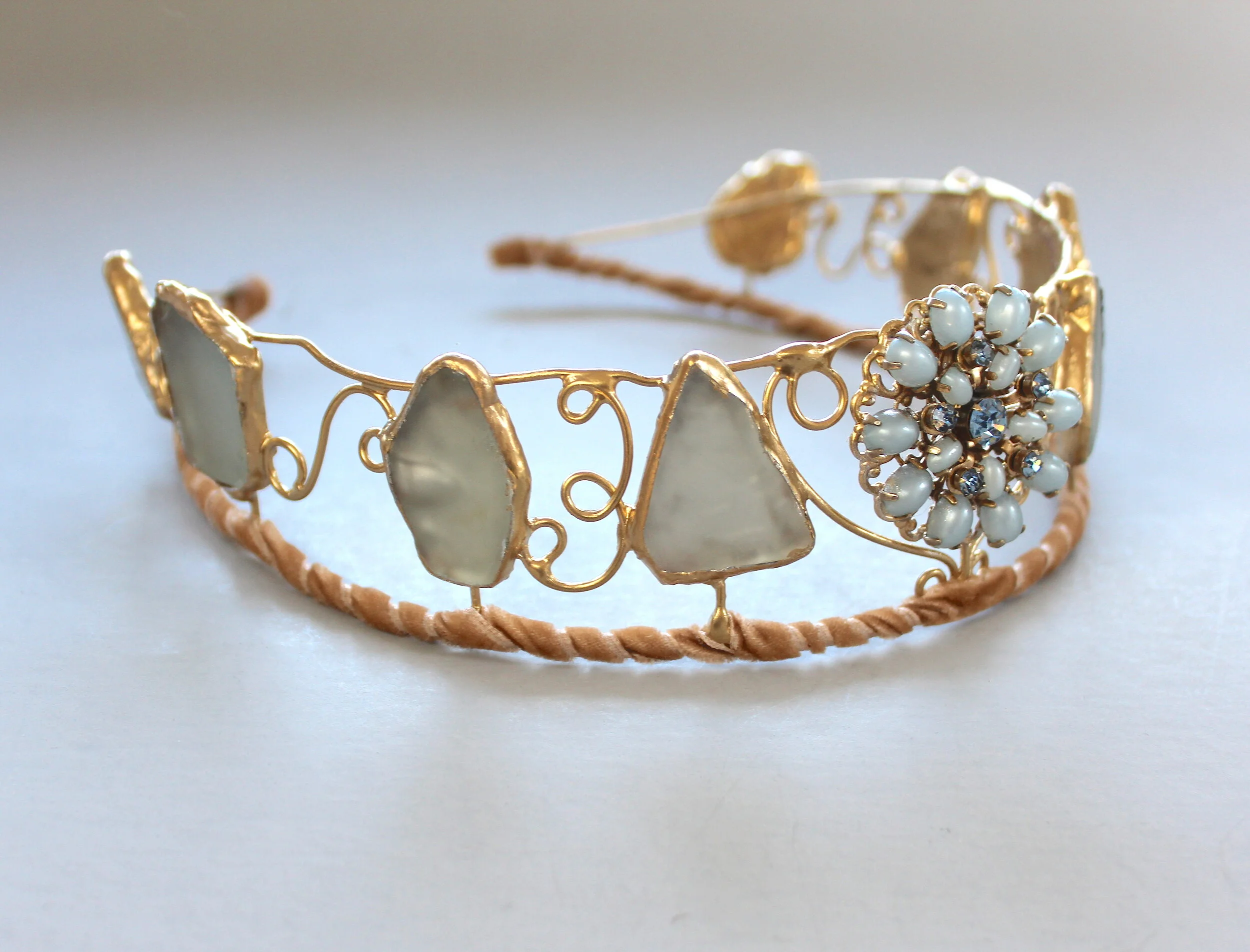 A decorative tiara with irregularly shaped white stones set in gold frames, featuring a cluster of small white pearls and blue gemstones on one side, and a twisted peach-colored band at the base.