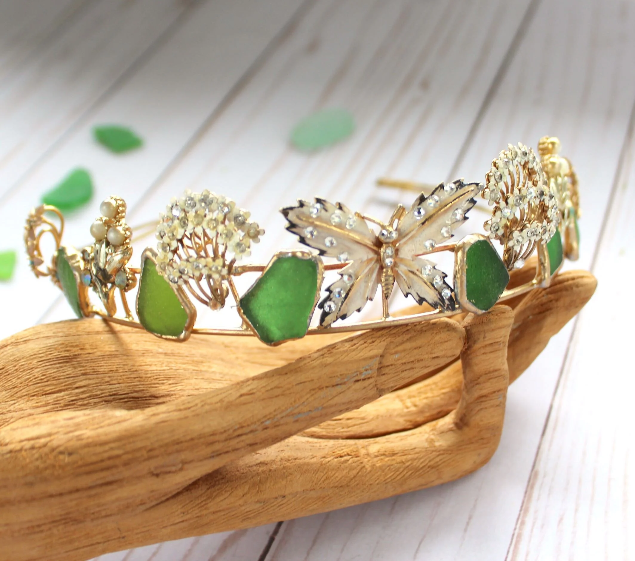 A decorative headband with butterfly and flower designs, featuring gold metal, white and green stone accents, and pearl details, resting on a wooden hand-shaped display against a white wooden background with green paper scattered around.
