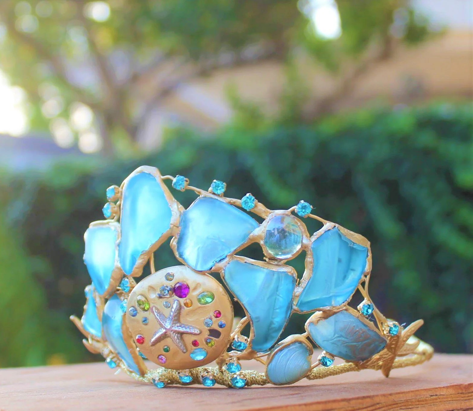 A decorative headband with blue shell pieces, colorful rhinestones, and a starfish charm, resting on a wooden surface outdoors.