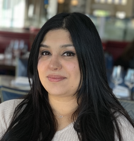 Smiling woman with shoulder-length black hair wearing a blue top, surrounded by green plants.
