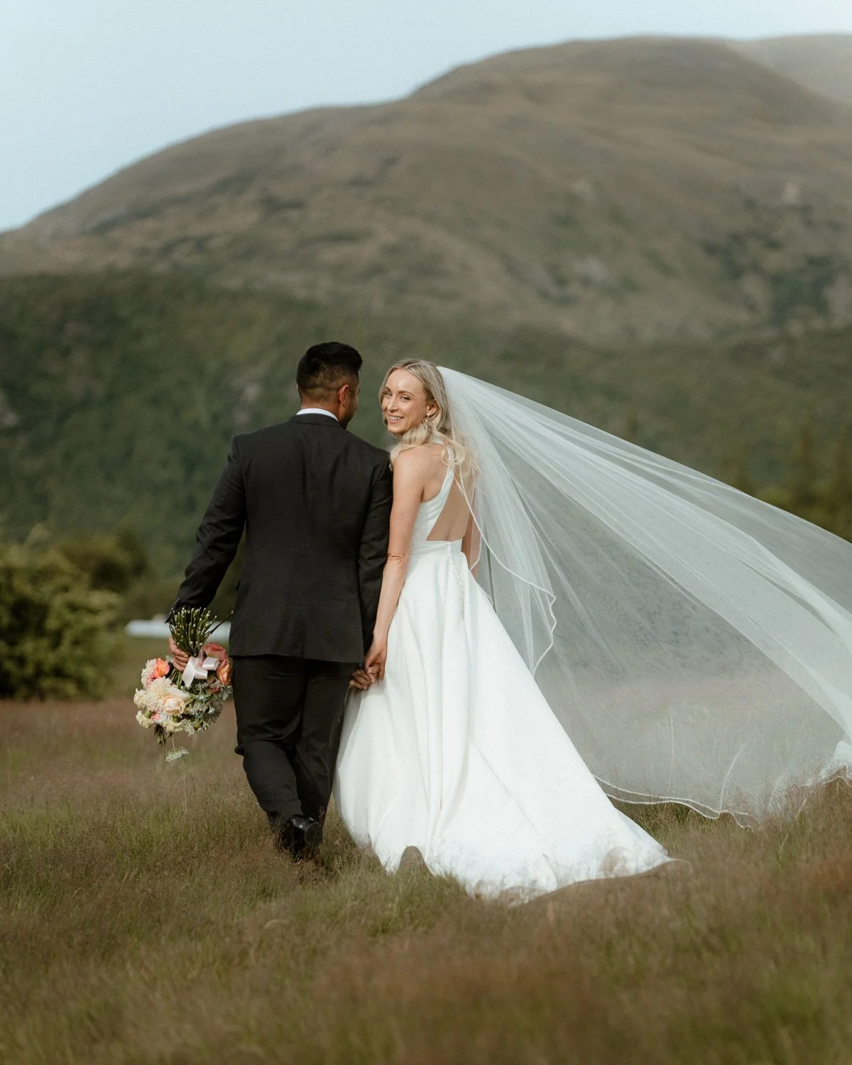 bride and groom walking toward Arrowtown from millbrook, summer grass and beautiful wedding veil blowing in the wind