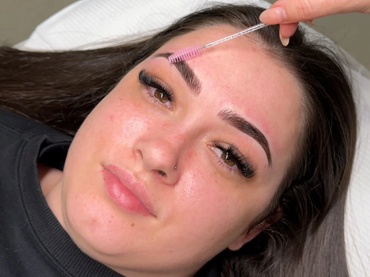 A woman lying down with her eyes open, receiving an eyebrow grooming treatment. An individual is using a spoolie brush to shape her eyebrows.