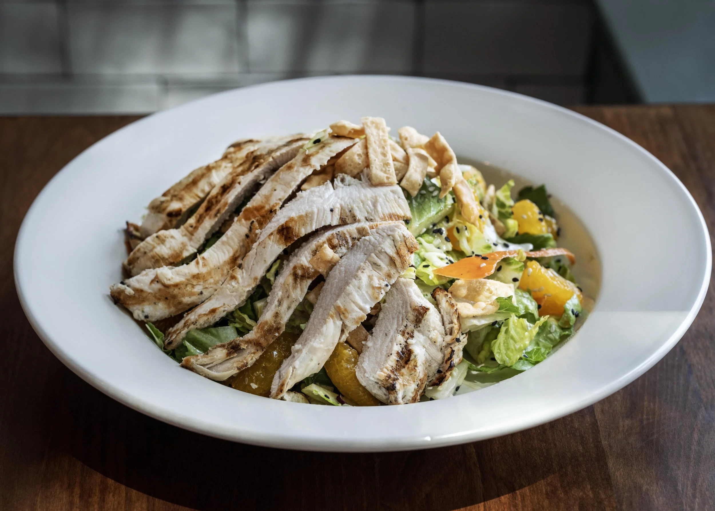 Grilled chicken sliced on top of a colorful mixed salad with vegetables and dressing, served in a white bowl on a wooden table.