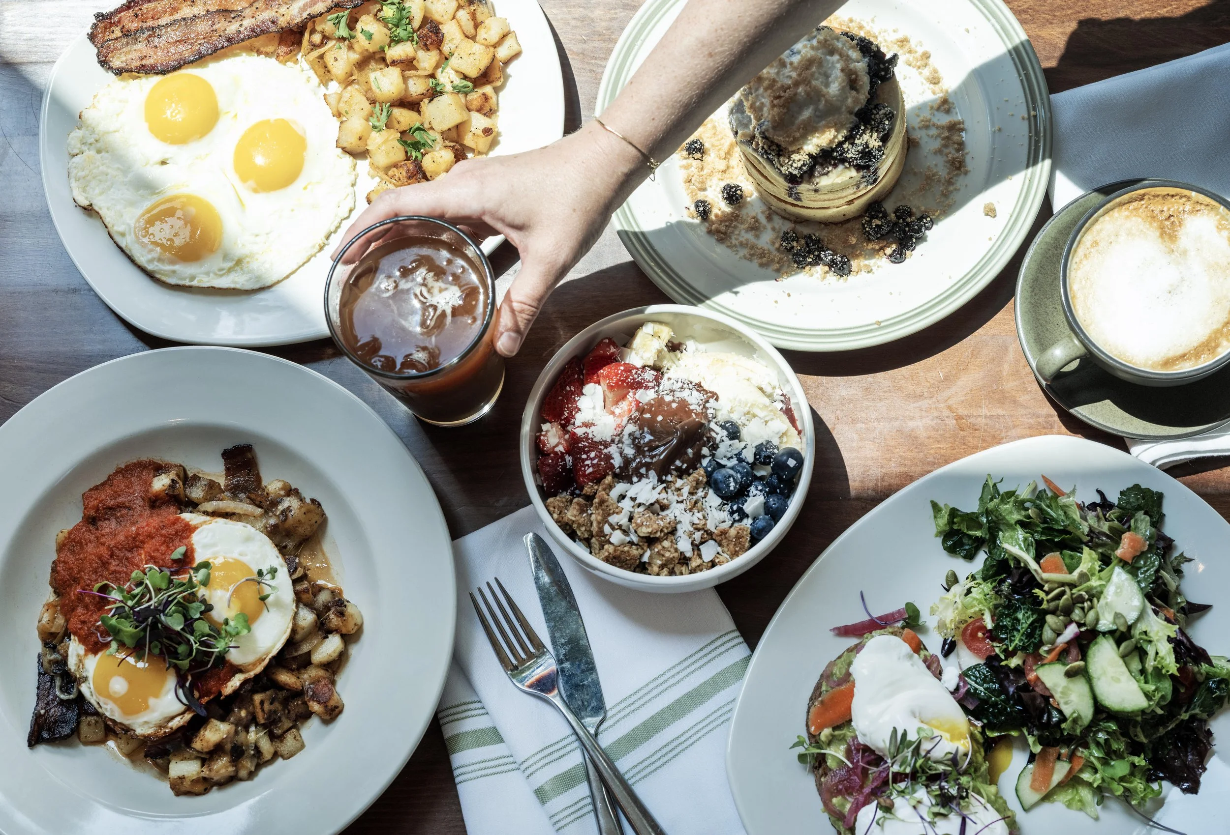 A breakfast spread including fried eggs with toast, hash browns, a bowl of fruit and yogurt, a plate of eggs with potatoes and tomato sauce, a mixed green salad with vegetables and a poached egg, a slice of cake with berries, and a glass of iced coff